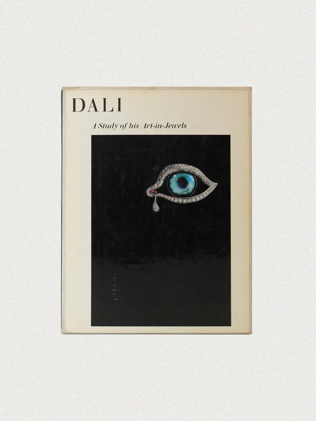 The book cover for "Dalí: A Study of his Art-in-Jewels" by Boga Avante Shop features a surreal eye-shaped jewelry design with a blue iris and teardrop on a black background, celebrating Dalí’s artistic legacy. The title is set in the top left corner.