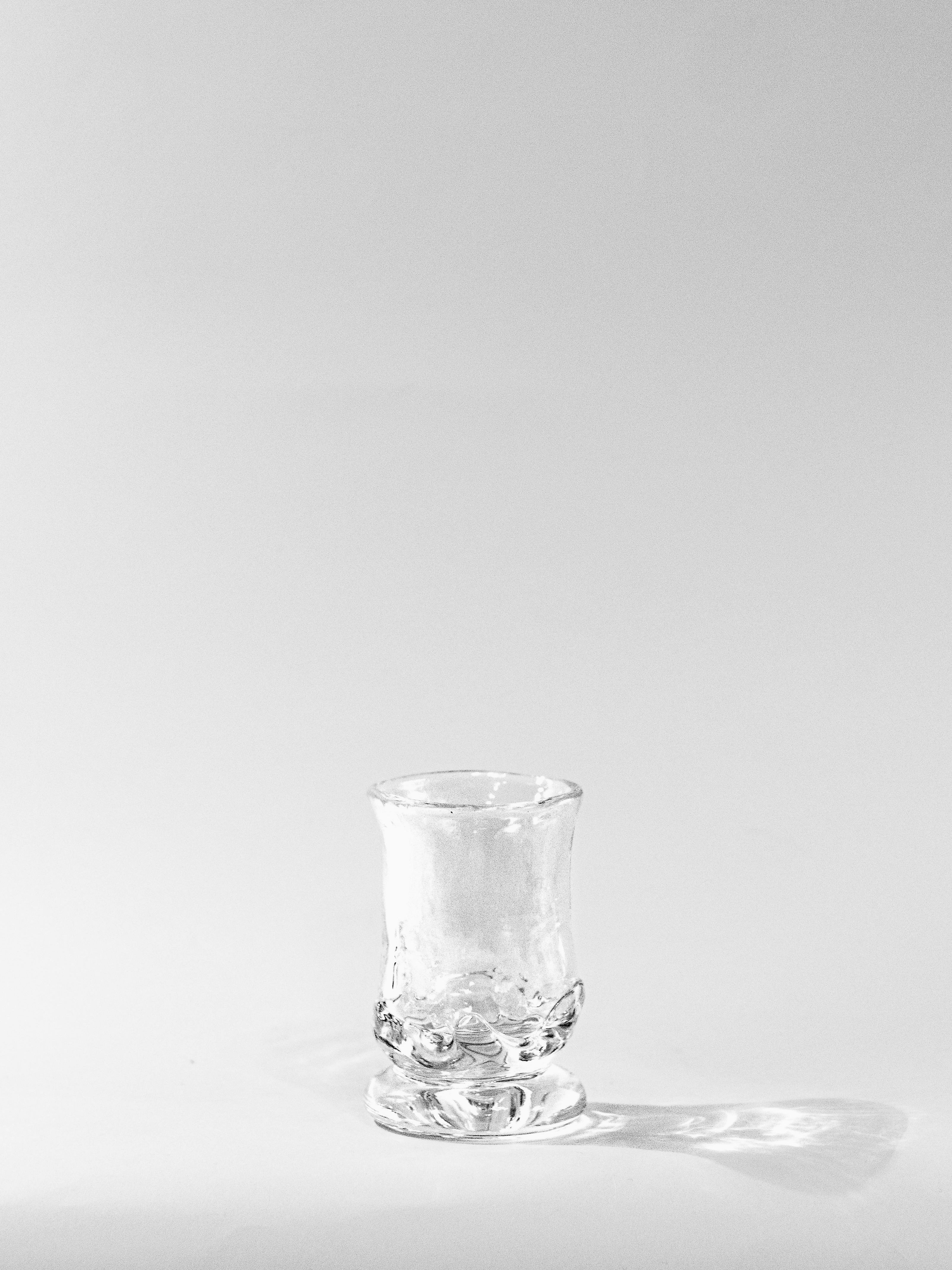 The SzkĆo Studio Narrow Goblet - Transparent, a small soda-lime glass with a textured surface, sits empty on a smooth, light-colored background, softly casting its shadow.