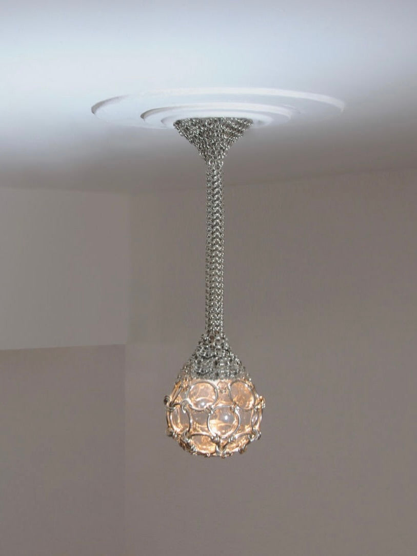 The Single-Pendant 'Chainmail Droplet' Chandelier by Panorammma is a modern, handmade fixture featuring a metallic chain and spherical shade with circular, translucent elements. It hangs elegantly from a white ceiling, creating a soft glow in the room.