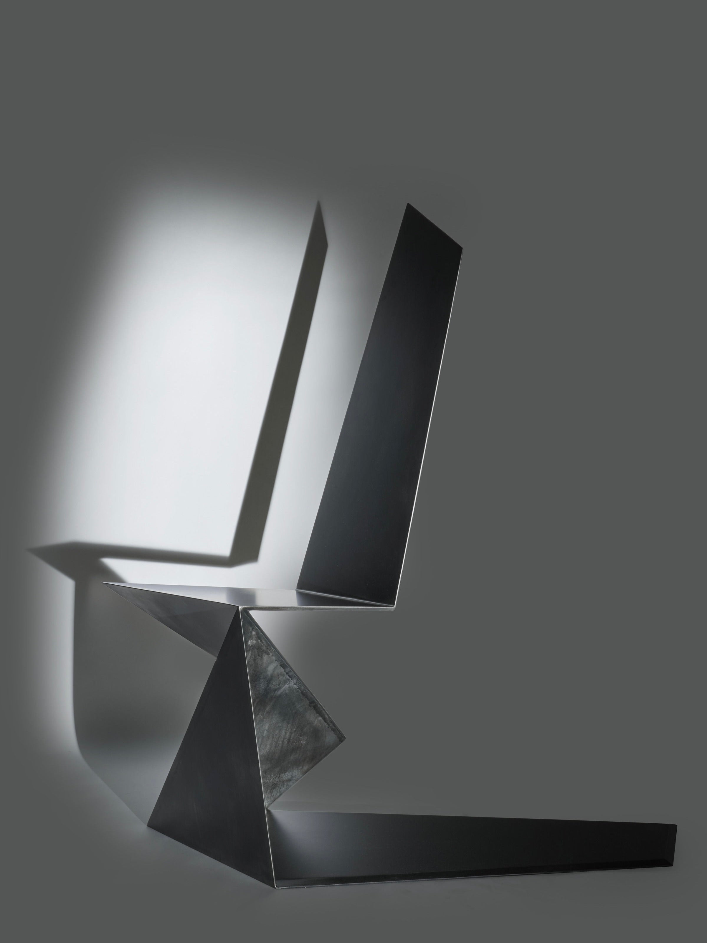 The Dark Side Chair by Orfeo Studio is a modern sculpture featuring sleek metal pieces with angular shapes and sharp edges, illuminated by soft lighting that casts chiaroscuro shadows on its muted gray background.