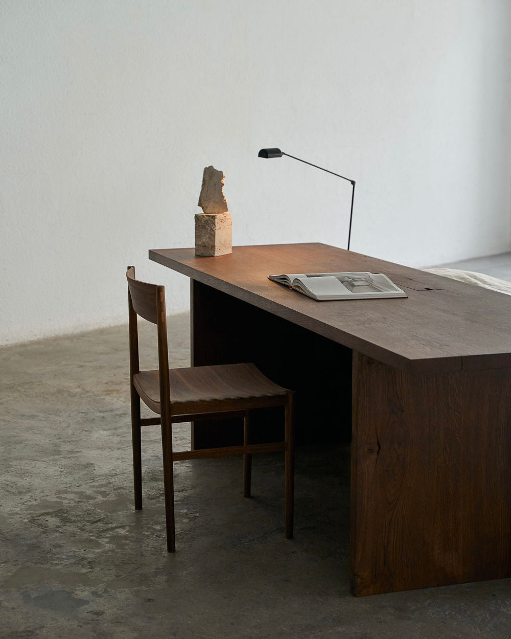 An Obstacles brand Ernest Desk paired with a simple chair sits in a sparsely decorated room, mirroring contemporary lifestyles. A modern lamp softly illuminates an open book on the desk, while a rough-textured stone sculpture rests elegantly on the left side of the writing desk.