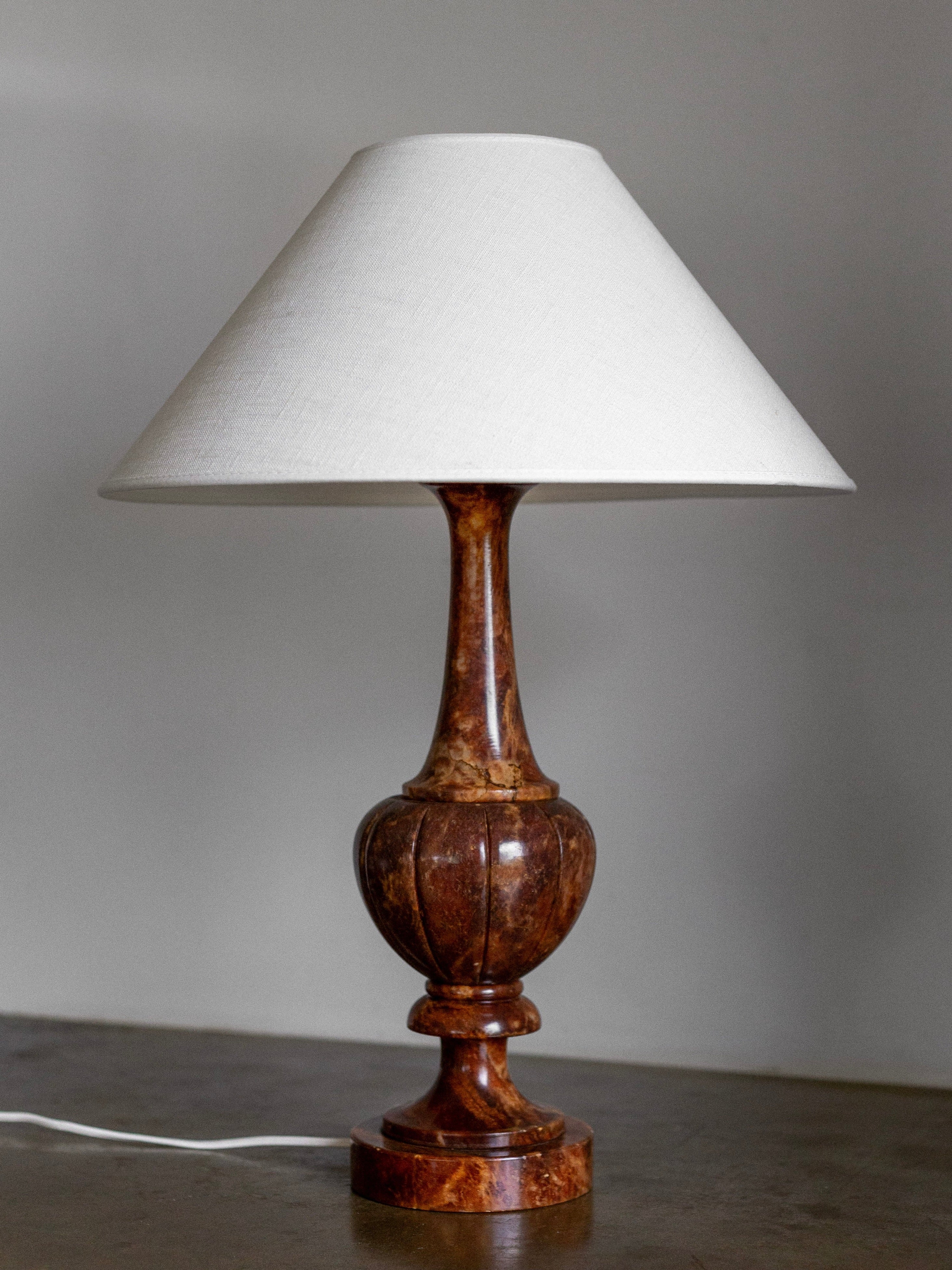 A Large Italian Alabaster Stone Lamp by Spigel, featuring a wooden base with a hand-carved design and topped with a white fabric lampshade. The lamp, exuding neoclassical style, is placed on a surface against a plain wall, with its white electrical cord visible at the base.