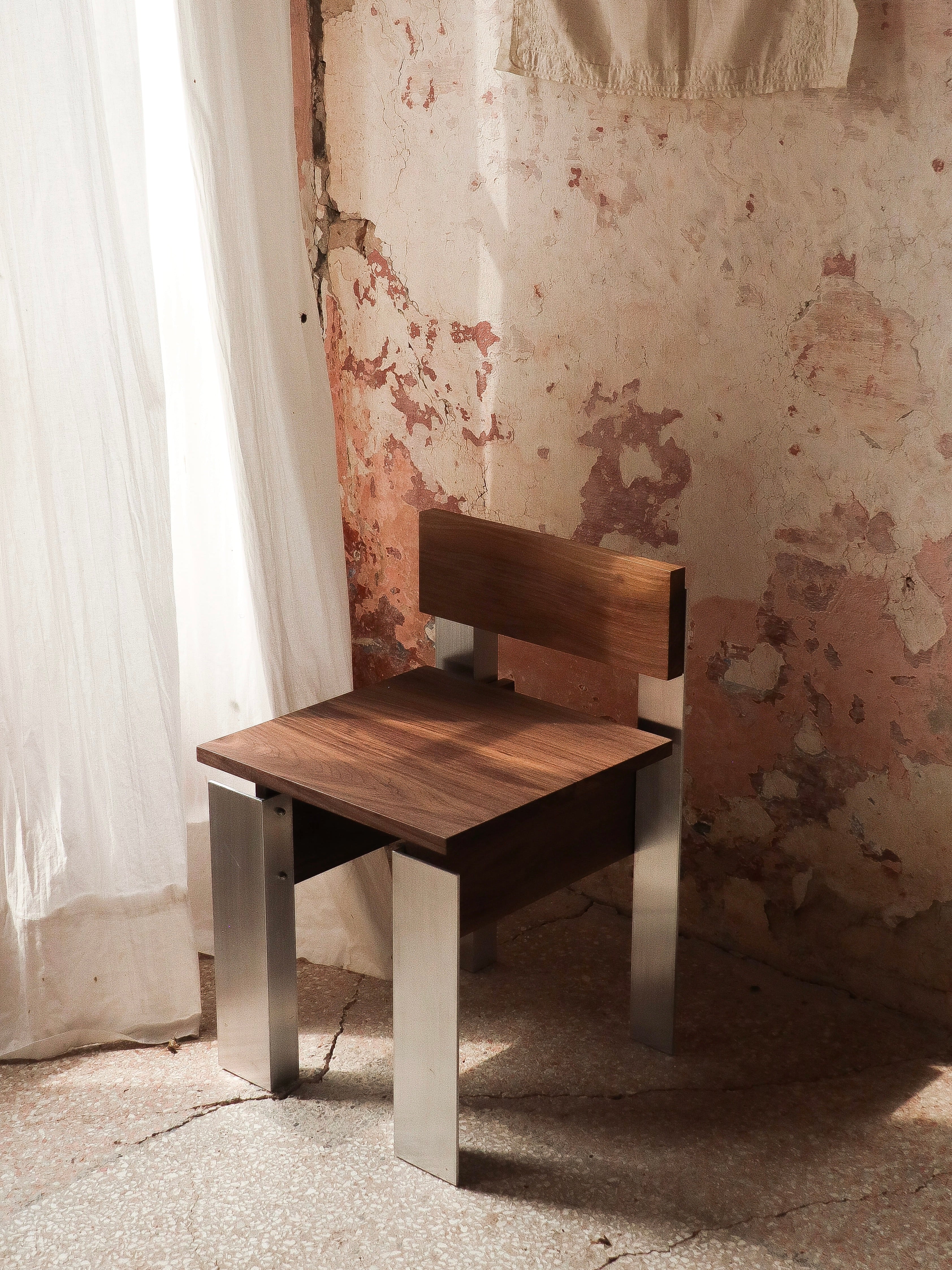 A Filippo Andrighetto Defender Chair, made of walnut with sleek aluminum legs, sits before a textured peeling wall. Soft natural light through a sheer curtain casts gentle shadows on the rustic stone floor.