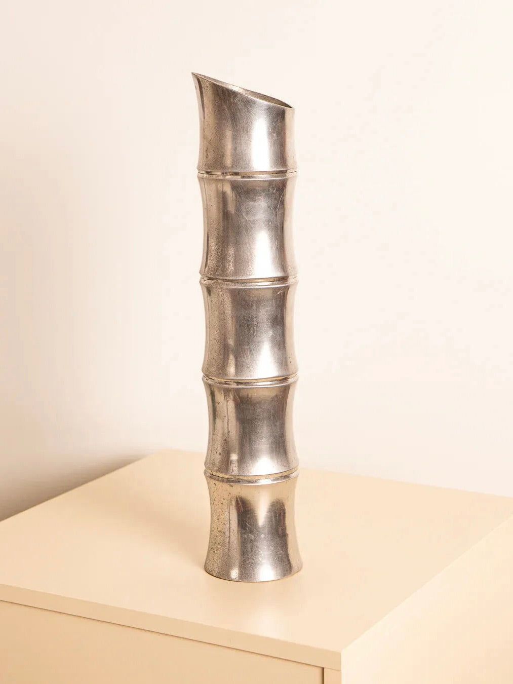 The Large Cast Aluminium Vase ‘Bambou’ 80's by Treaptyque, a tall silver metallic piece, takes the form of a bamboo stalk with multiple segments. It exudes vintage charm while standing on a light-colored wooden surface against a plain beige background.