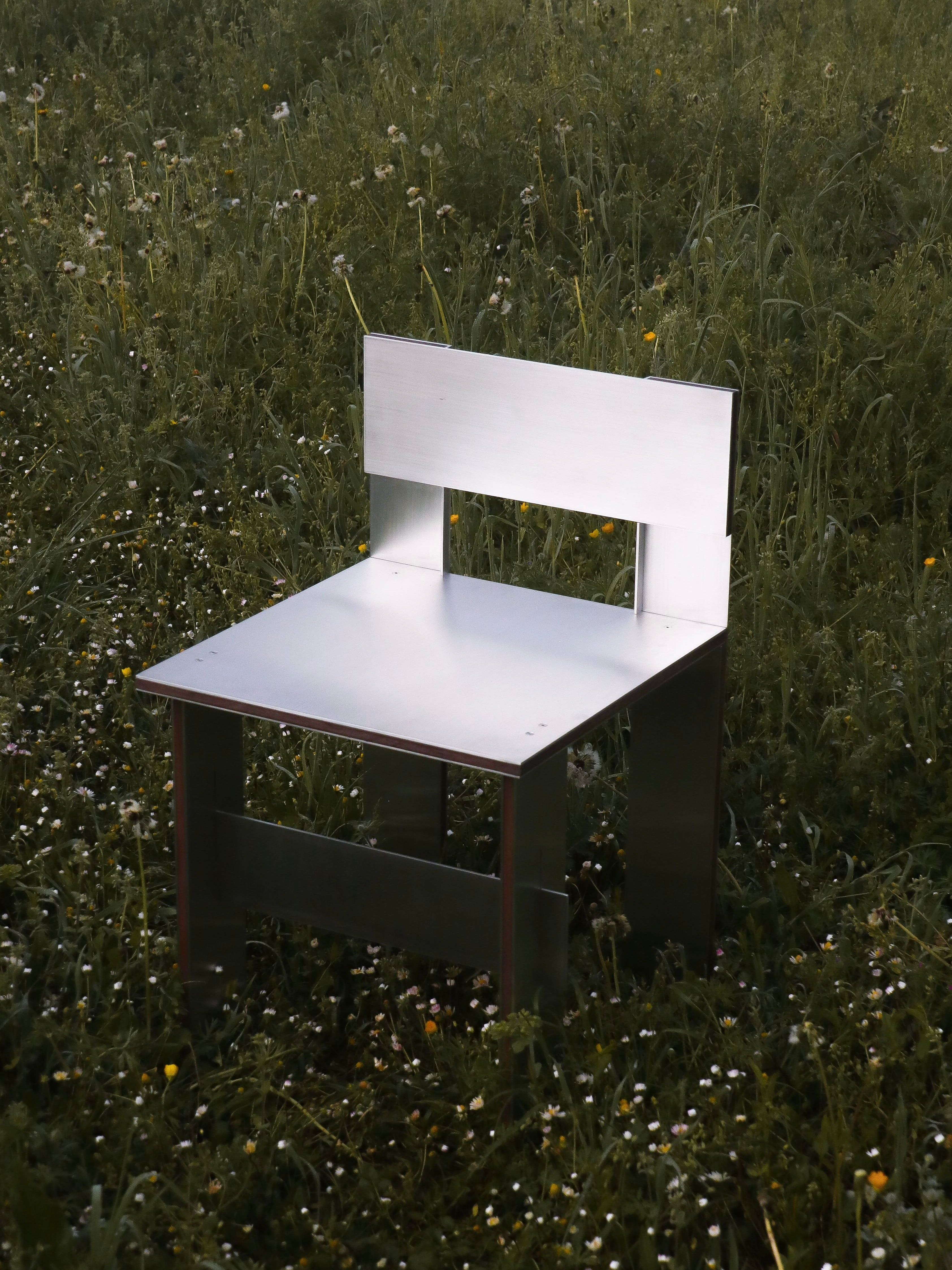 The Sandwich Chair by Filippo Andrighetto, featuring a sleek minimalist design with a rectangular backrest in walnut and aluminum, sits on a grassy field adorned with small flowers and dandelions, creating a modern contrast to the vibrant natural surroundings.