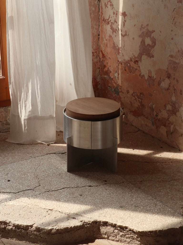The Lunar Side Table by Filippo Andrighetto, with its round aluminum frame, is positioned on a cracked floor in a sunlit room. The walls display a rustic, peeling texture, and sheer white curtains hang by a wooden window, letting in gentle natural light.