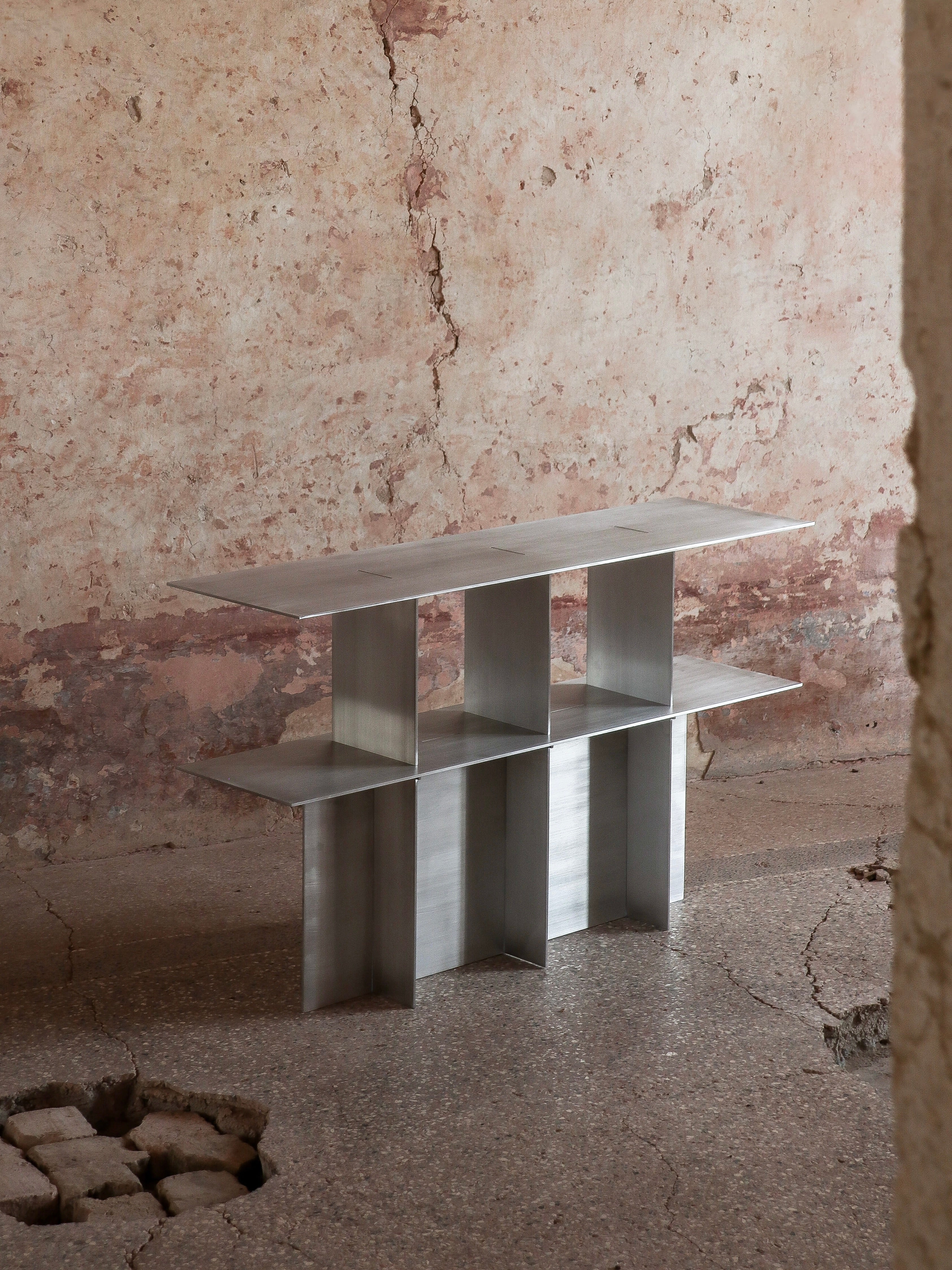 In an unfinished room with cracked concrete walls and floors, the Lamina Console by Filippo Andrighetto stands out. Its minimalist design features brushed aluminum surfaces supported by four vertical panels, embodying a geometric and industrial aesthetic.