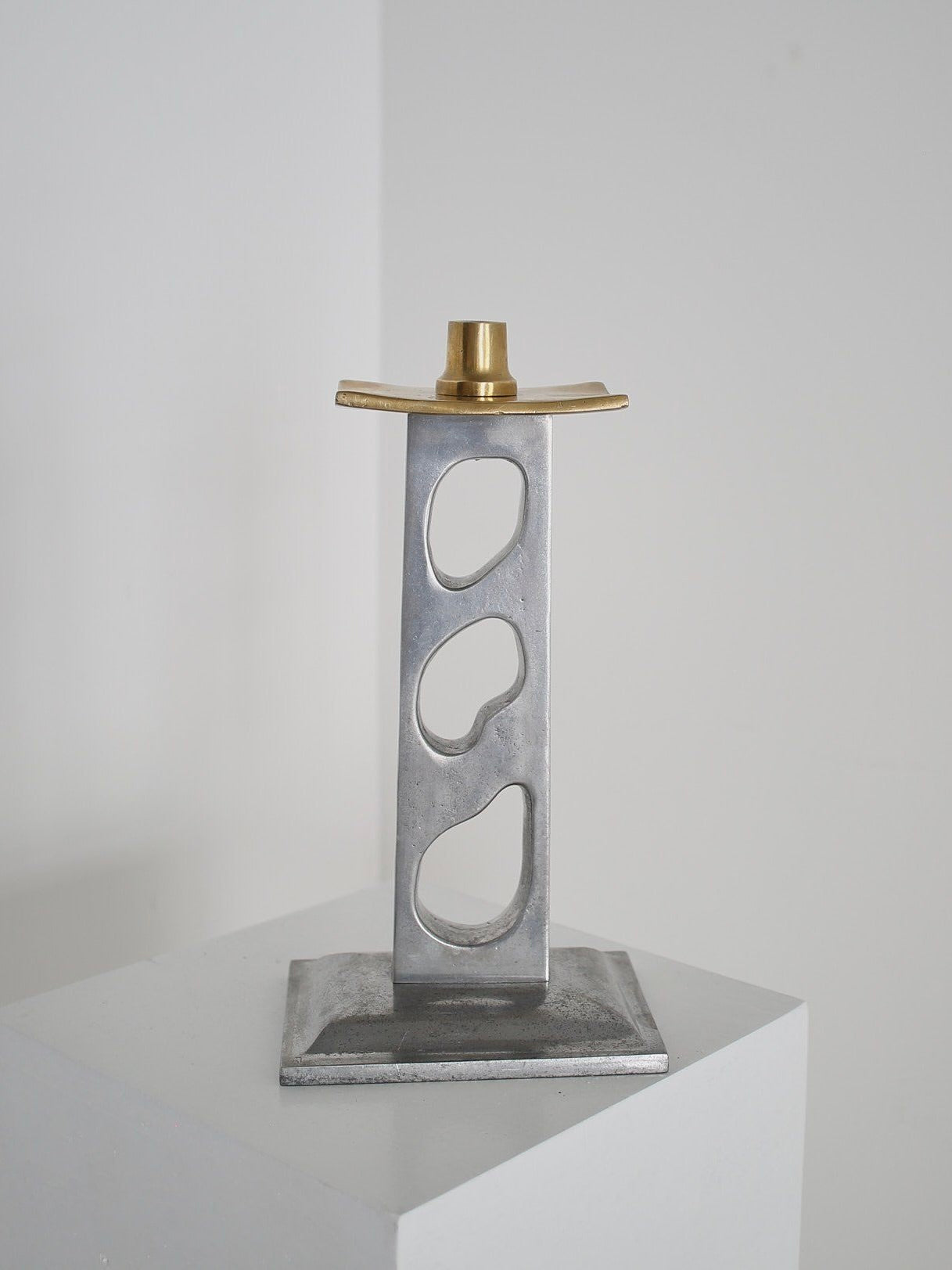The David Marshall Candleholder by Ancien et Jolie is an abstract minimalist sculpture made of cast aluminum, resembling a 1970s candlestick. It has a tall rectangular shape with three irregular cutouts and a brass-colored top, exuding timeless elegance against a plain white background.