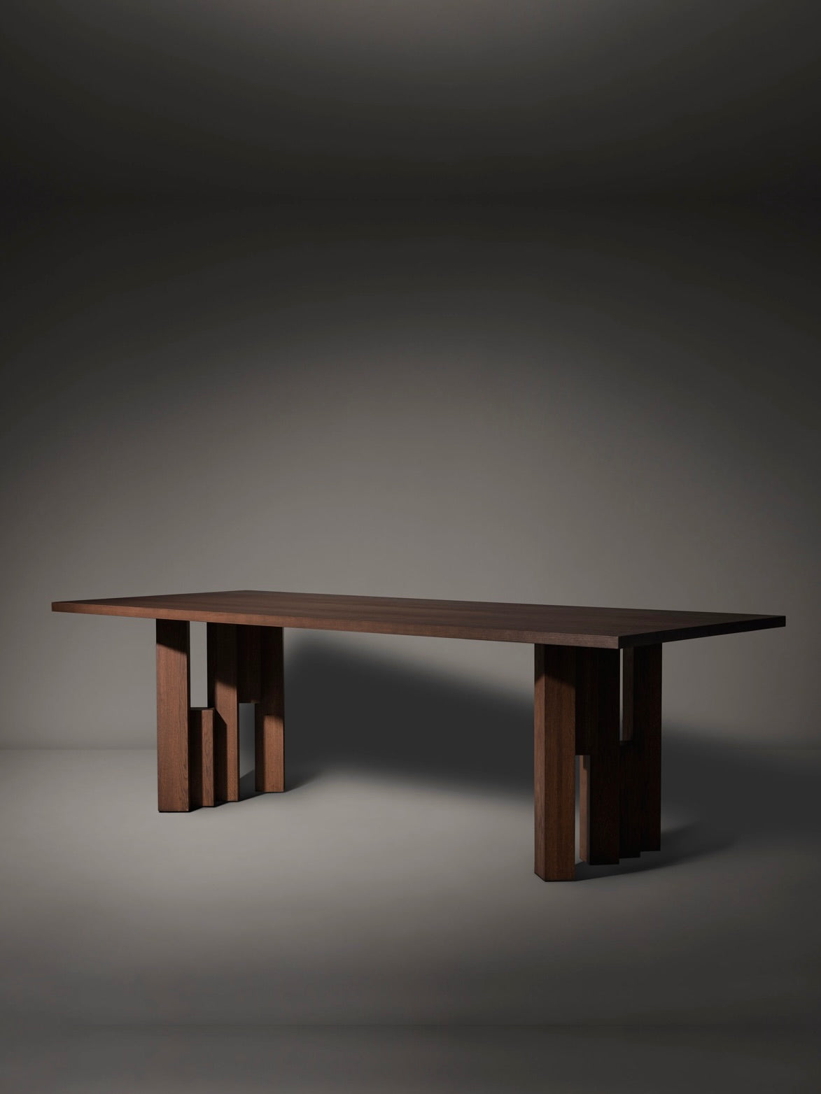 The Mokko Fenestra Table, made from European oak, features a modern design with a flat rectangular top and unique legs with vertical and horizontal slats. It elegantly stands on a smooth gray surface against a dark background.