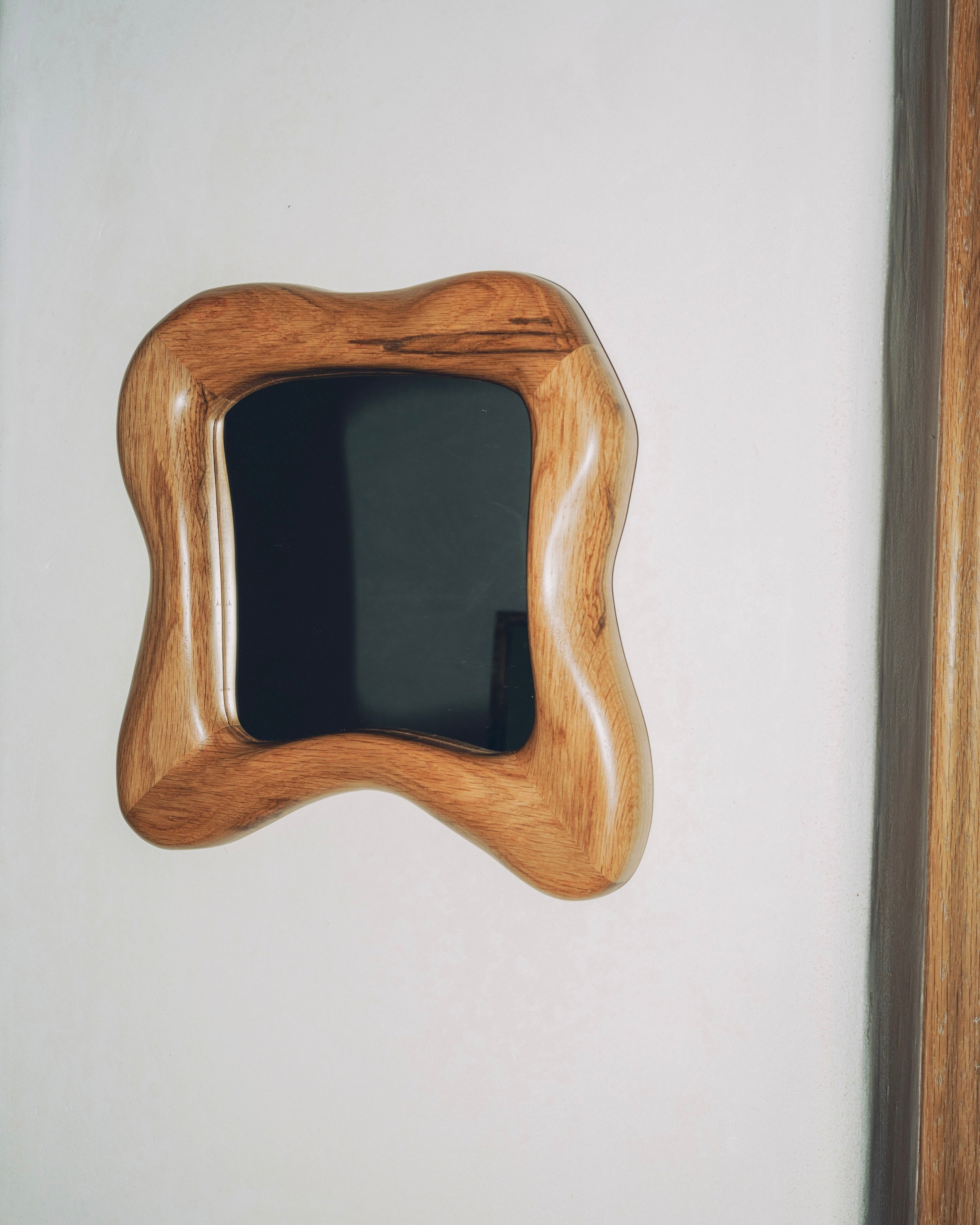 A compact, square Minho Mirror Oak by Project 213A, featuring a wavy solid wood frame handmade in Portugal, hangs on a plain white wall near a wooden edge.