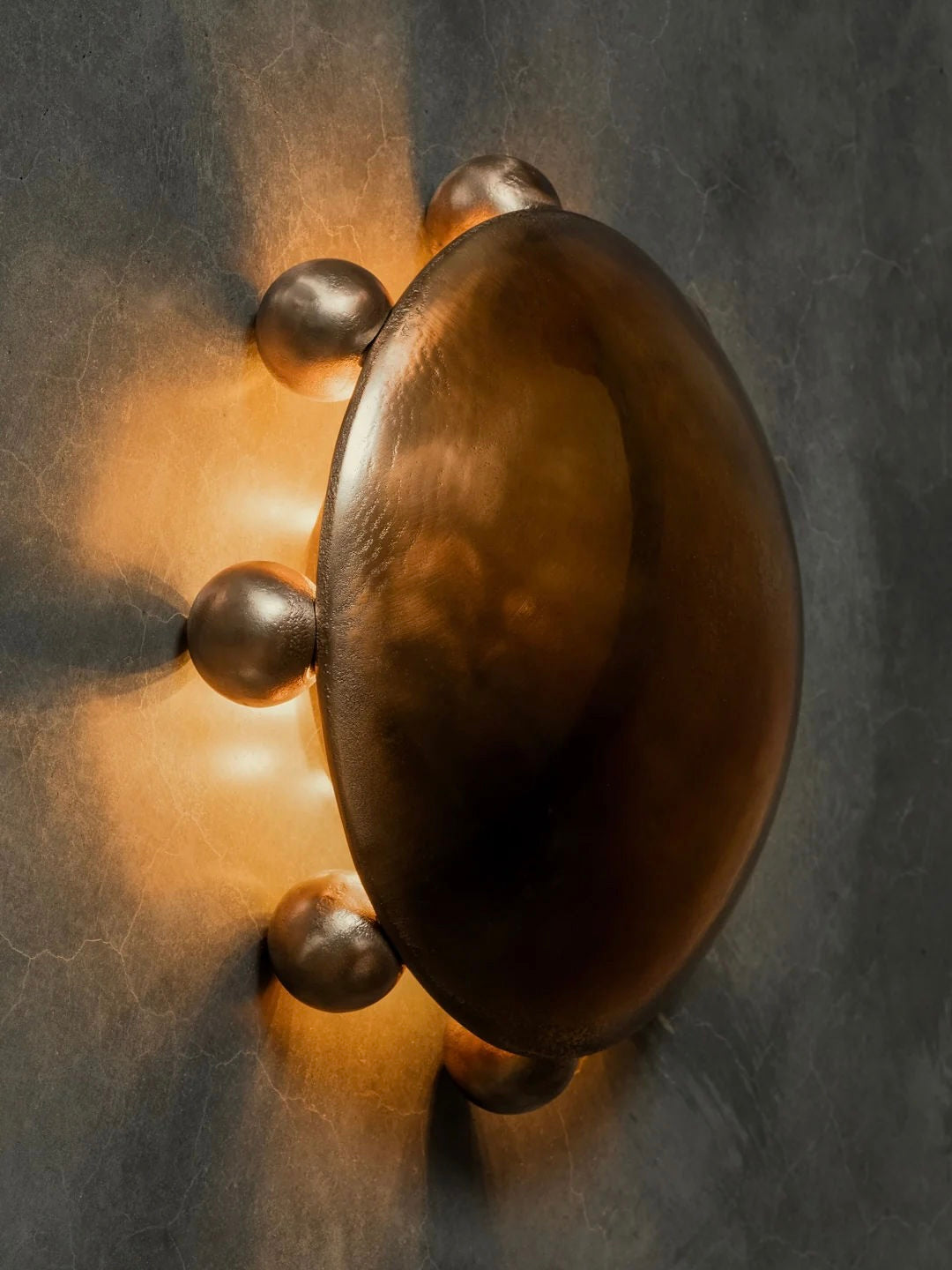 The Kaytar "ALTER" Sconce in bronze, a sand-cast oval disc with bulbous protrusions, emits a warm glow. Mounted on a textured gray wall, it creates a starburst pattern, adding alchemy to the space.