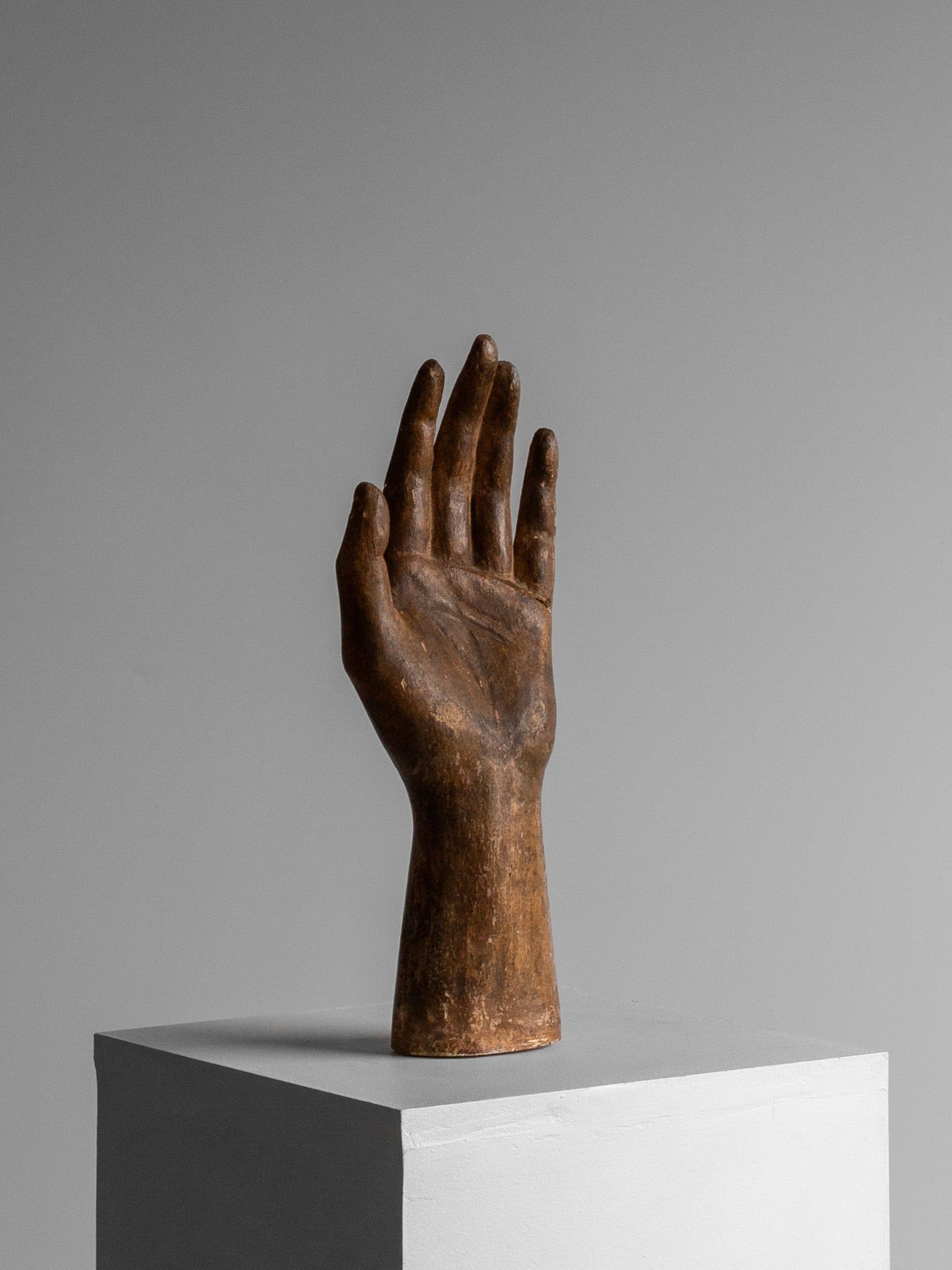Crafted with exquisite detail, the Spigel Antique Wooden Hand Sculpture features fingers slightly spread, elegantly showcased as a decorative accent on a white pedestal against a plain gray background.