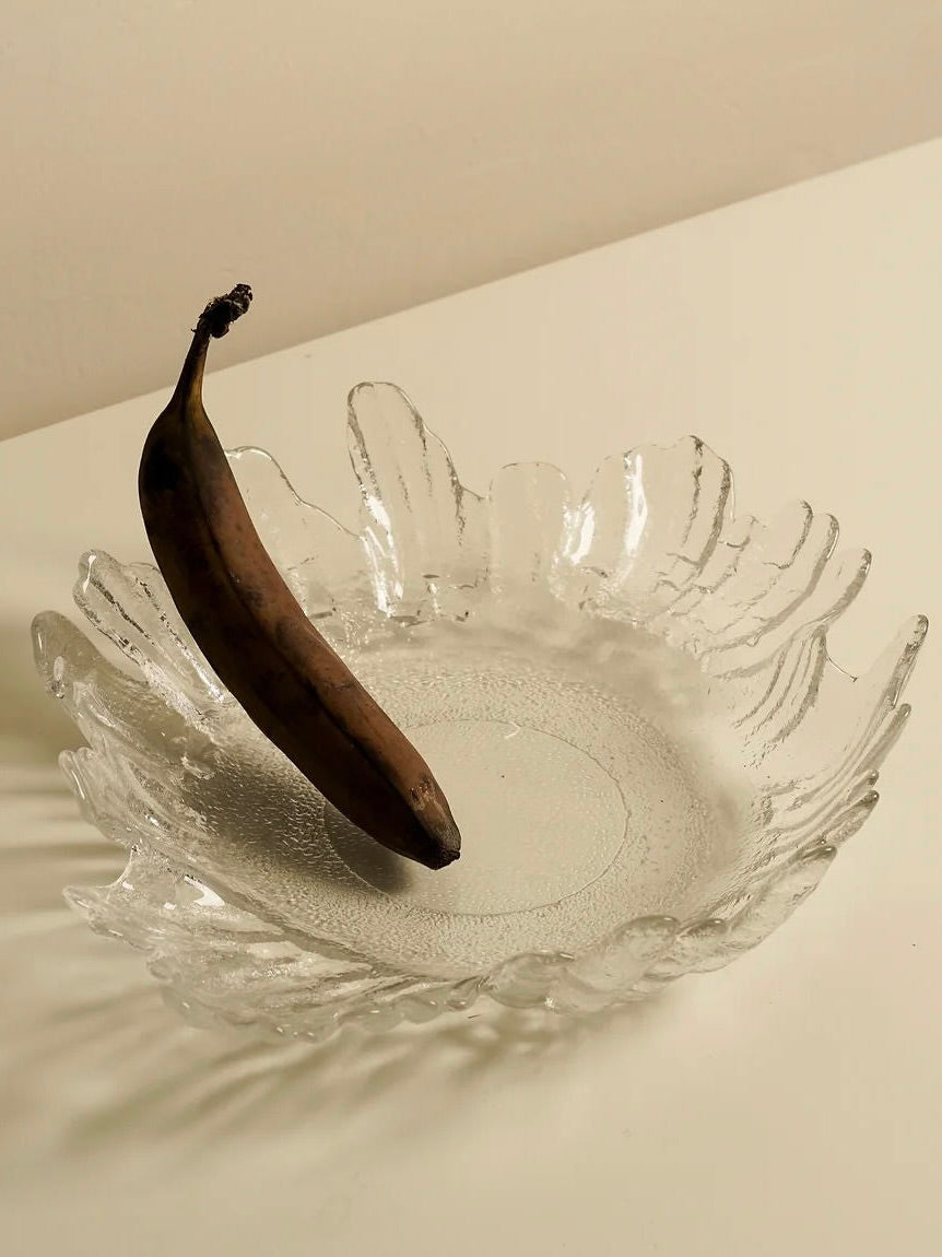 A single overripe banana with a dark peel rests inside an elegant Lindshammer Bowl from Les Objets Amsterdam, featuring a scalloped rim and placed on a light-colored surface. Shadows cast by the bowl and banana add depth to the minimalistic composition, reminiscent of Mediterranean homeware aesthetics.
