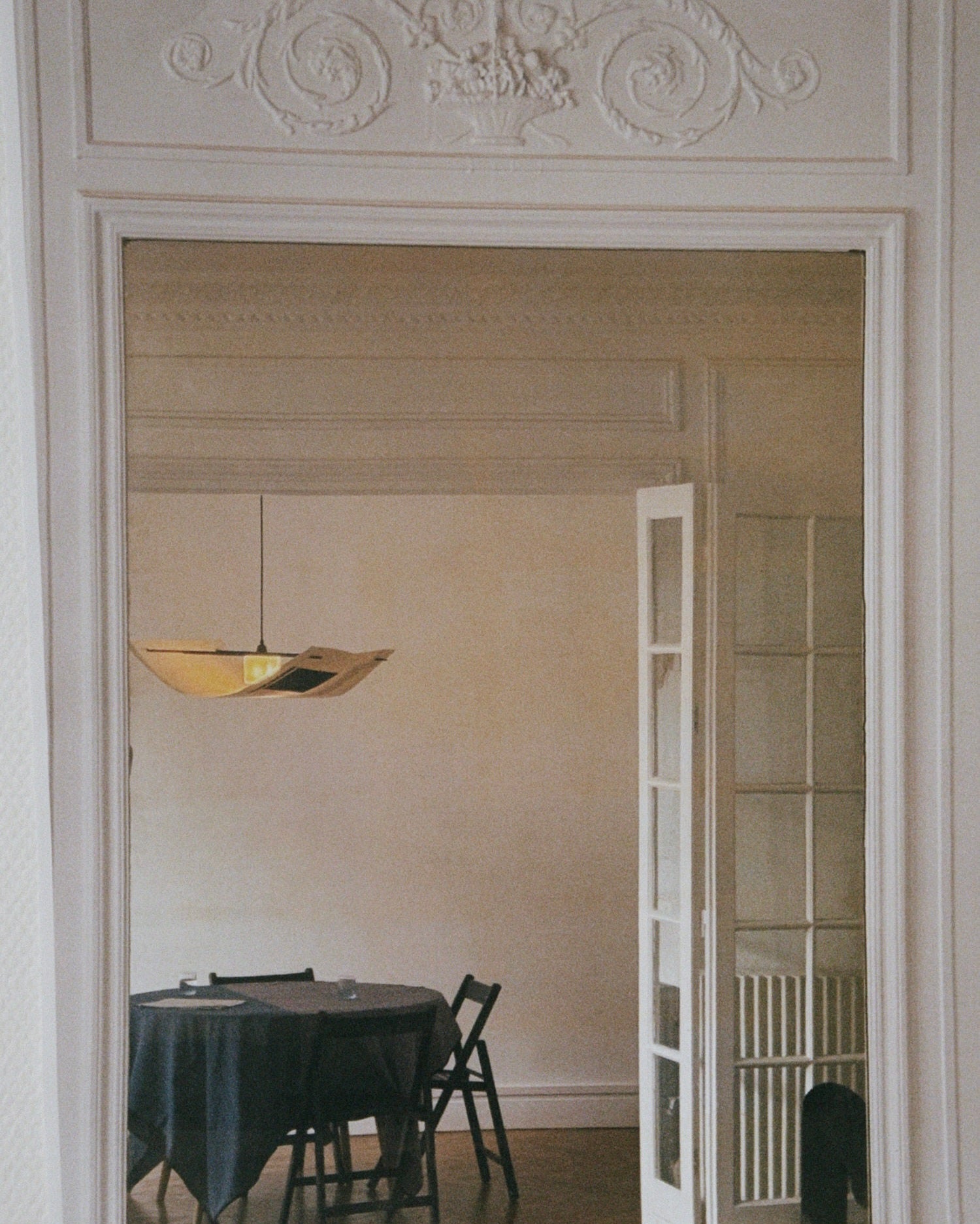 A mirror reflects a dining room featuring a handmade Sail Lamp – Rice Field by TEGET above a round, dark-clothed table. The room includes ornate molding, partially open French doors, minimalist decor, and unique paper collage art.