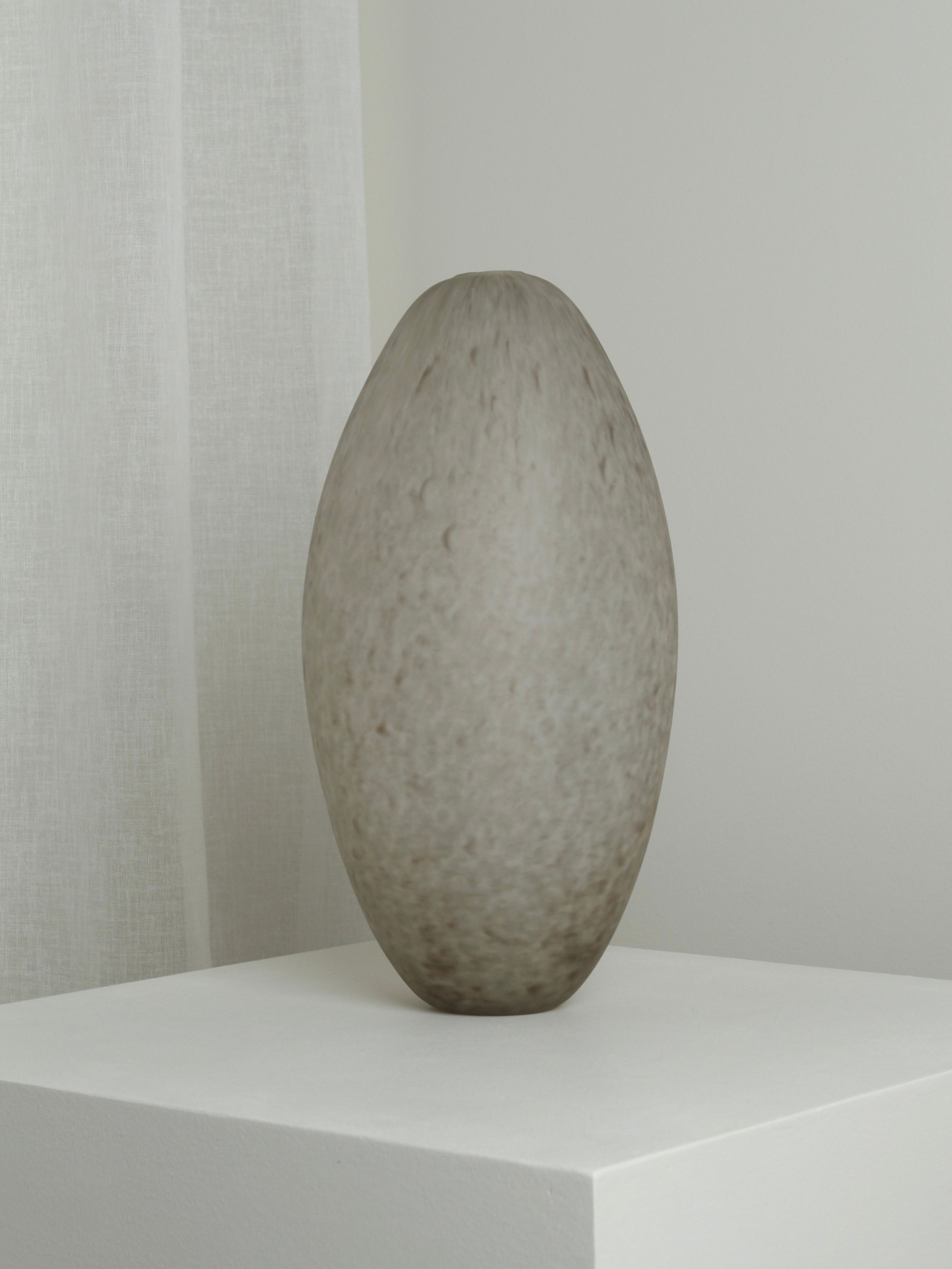 A large, egg-shaped XLarge Vase in Feathered Taupe by Verre d'Onge, featuring a textured design, stands on a white pedestal. This mouth-blown piece is set against a light gray wall with sheer white curtains, exuding a minimalist and modern aesthetic.
