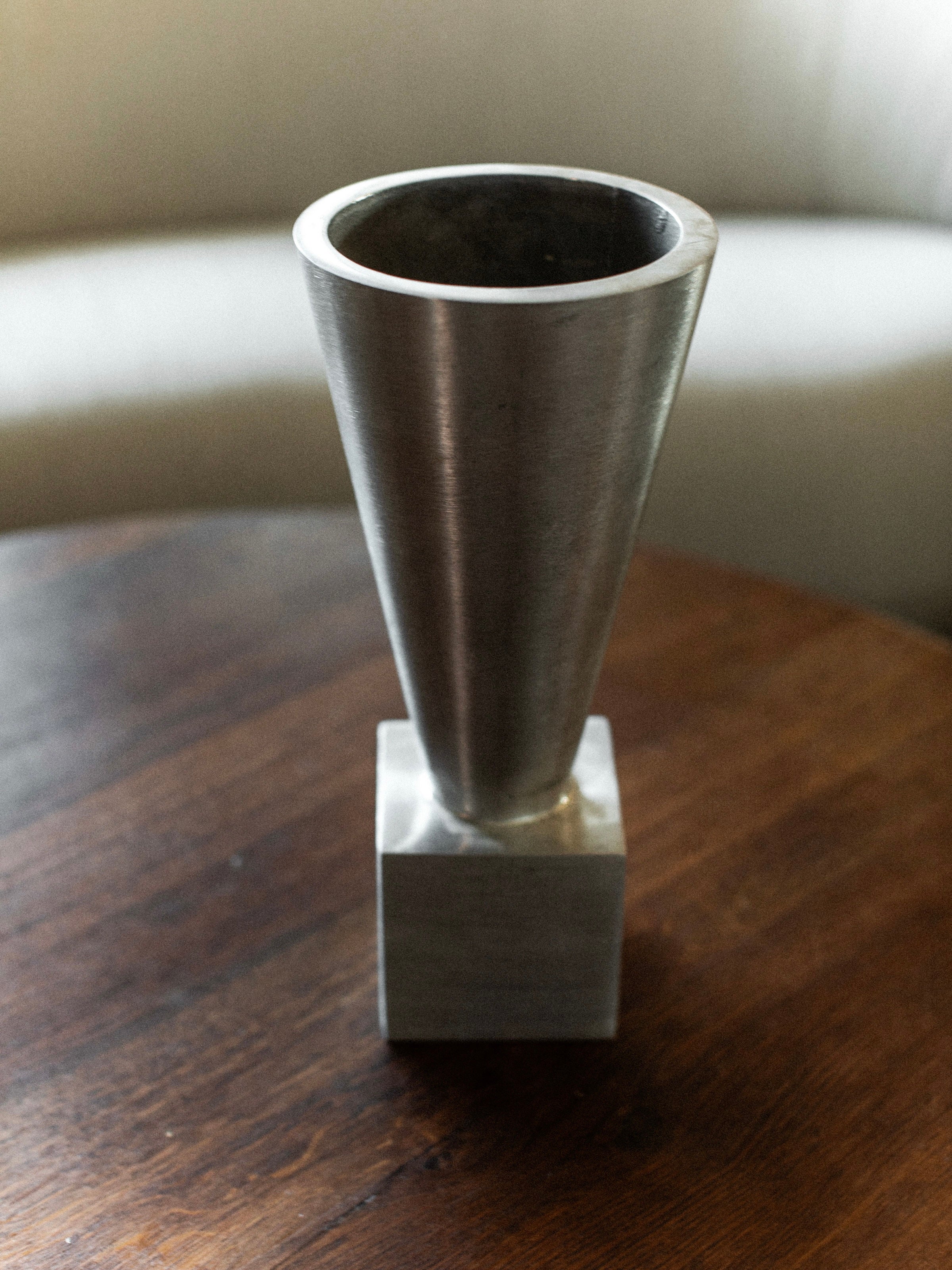 The 70's Aluminum Cast Vase by Out For Lunch, featuring a minimalist cone-shaped form and wide opening in brushed aluminum, sits elegantly on a wooden table. Its modern design provides a striking contrast to the soft, light-colored couch in the background.