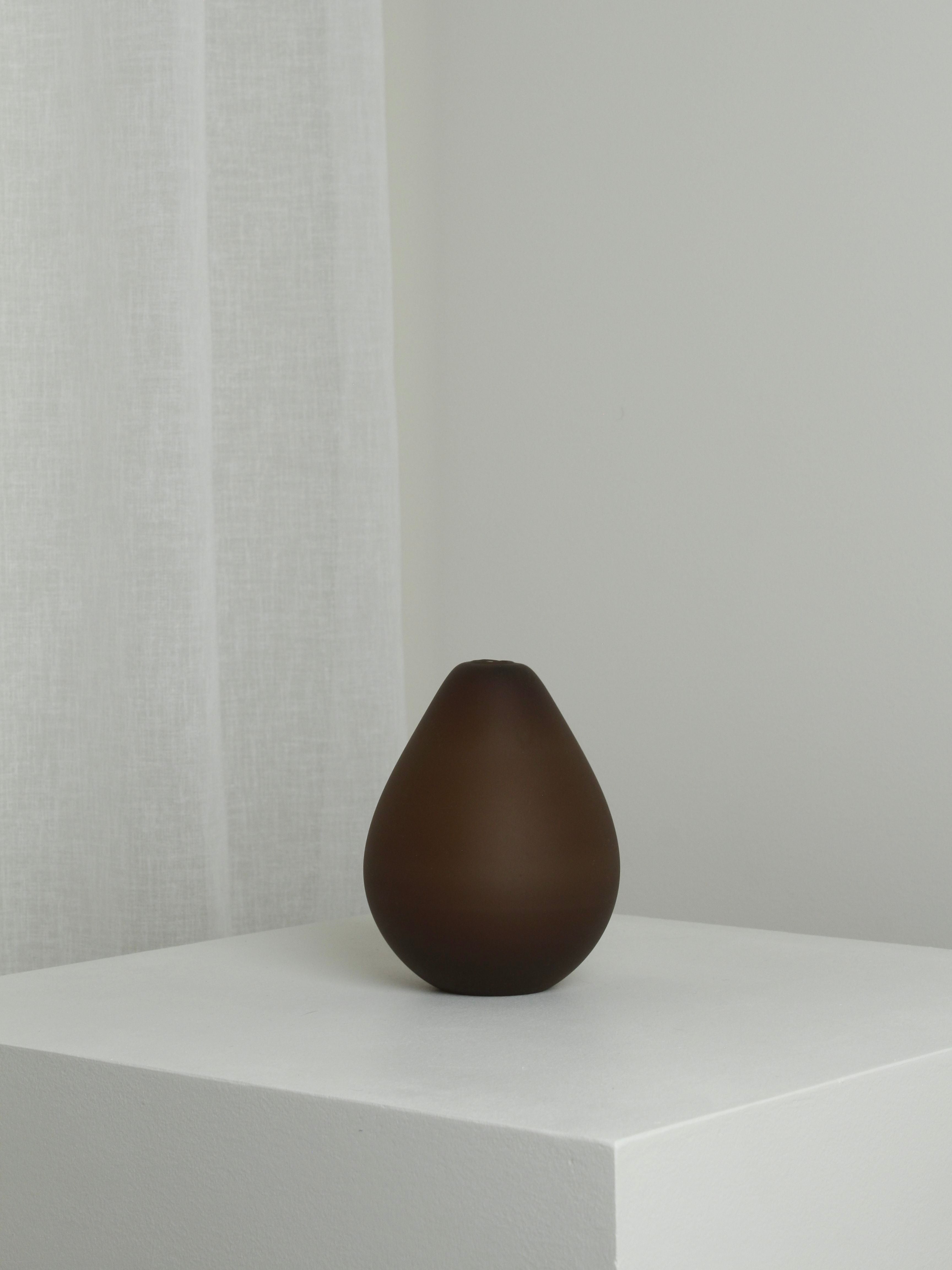 A minimalist image featuring the Medium Vase in Chocolate by Verre d'Onge, a mouth-blown teardrop-shaped vase, placed on a white pedestal. The background has a soft white curtain and a light gray wall, creating a calm and simple aesthetic.