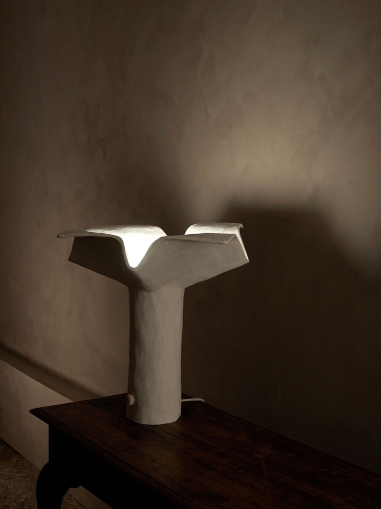 The 'La Fleur' Table Lamp by Studio Tristan Kallas features a unique white ceramic design with an abstract flower-like top, casting floral silhouettes and soft light on a dark wooden table, enhancing the textured neutral-toned wall with a minimalist artistic vibe.