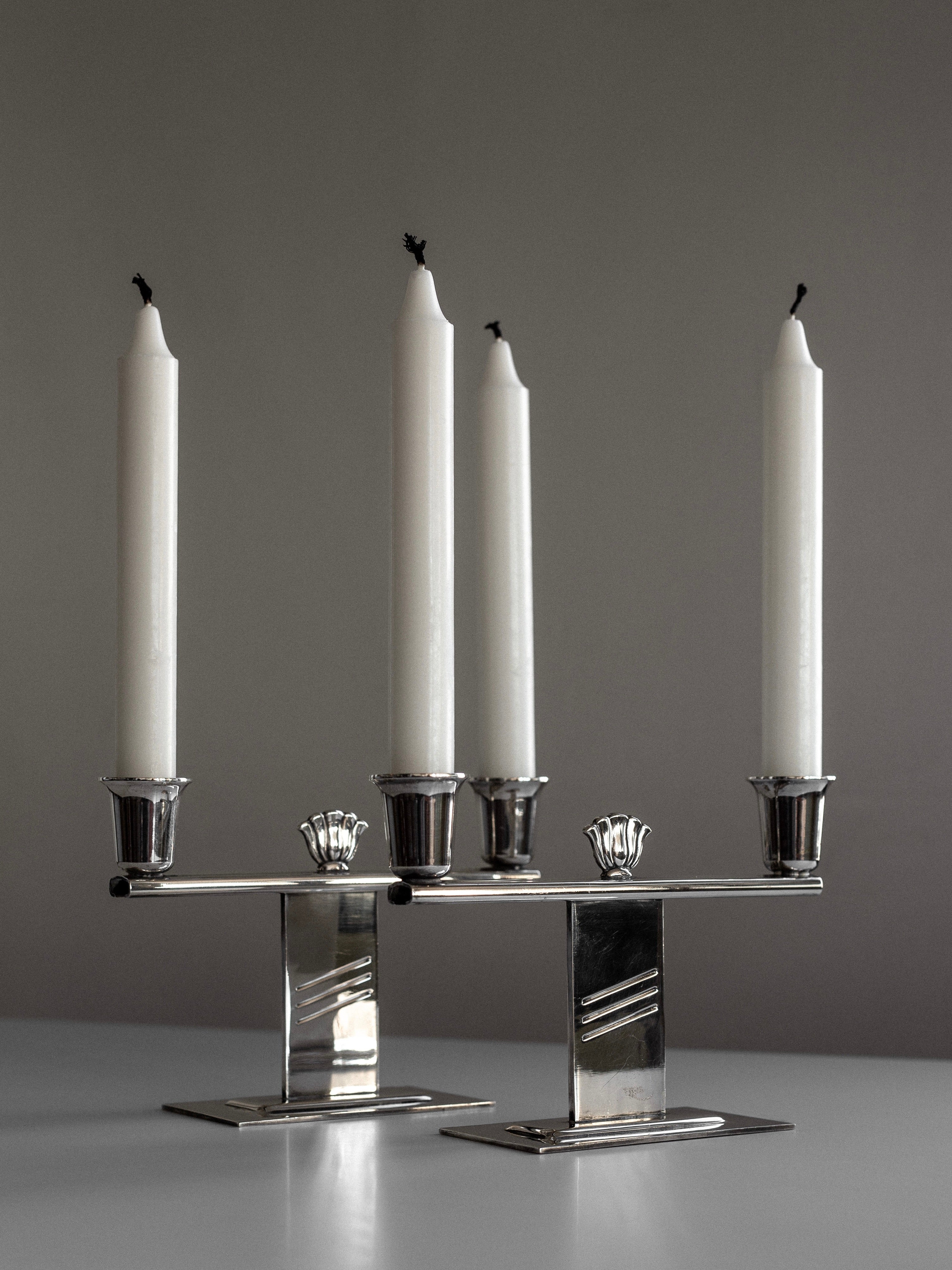 A pair of Spigel silver-plated Art Deco candle holders stands on a light surface against a neutral background. Showcasing Scandinavian craftsmanship, they feature a sleek design with decorative elements and hold four unlit white candles.