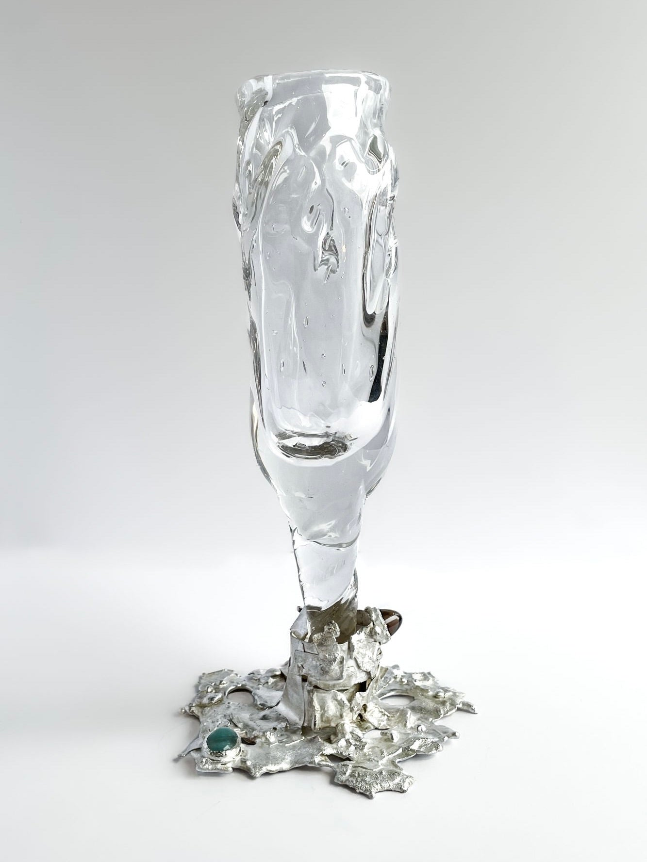 The Jewelled Goblet no. 6 by Lindsey Fontijn, offered by Szkło Studio, is a sculptural glass artwork featuring a wavy, twisted form that extends upward from a base adorned with star shapes and natural gemstones. This piece showcases an abstract, modern design with a clear, smooth hand-blown glass surface and a metallic foundation.