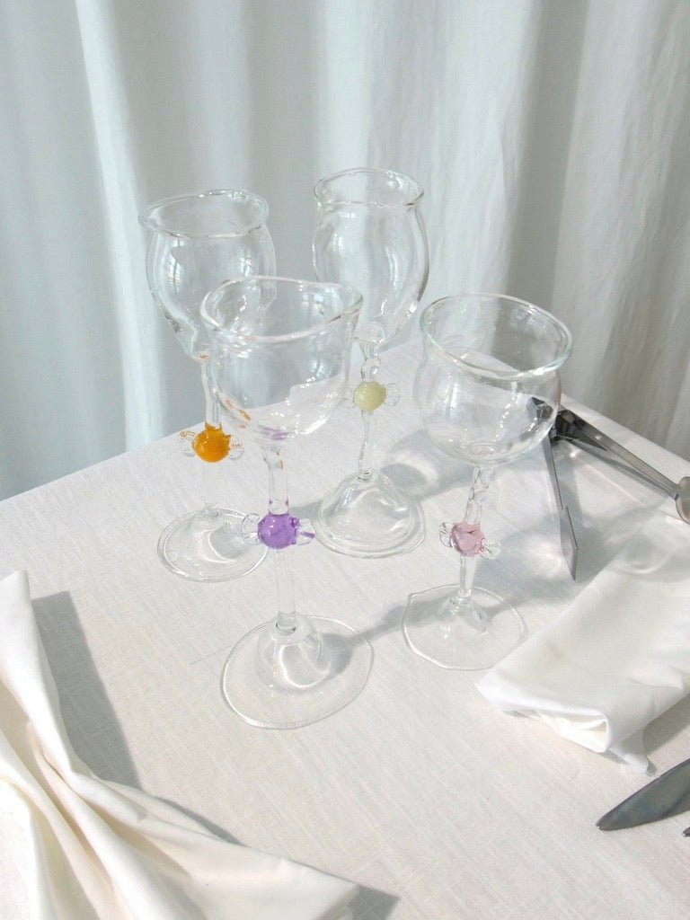 Four Mouth Blown 'Candy' Wine Glasses by Vivian Alarcón, each featuring colorful stem accents, are arranged on a white tablecloth beside folded napkins and cutlery, with a white curtain in the background.