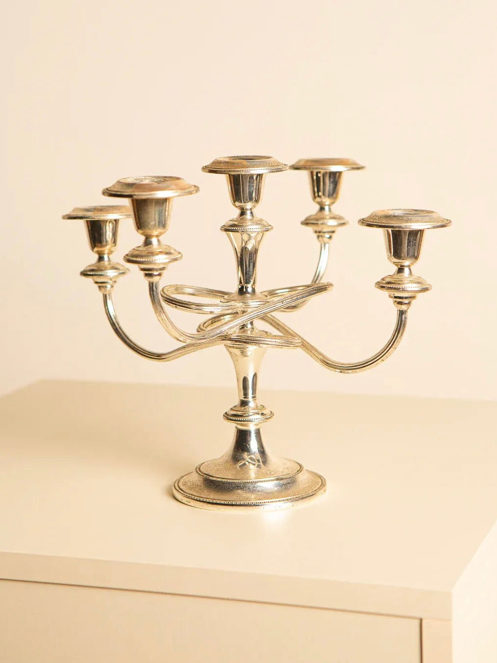 The "Medusa" 60's Italian Candlestick by Treaptyque, a silver candelabra with five candle holders, sits on a cream-colored surface against a light beige background. Its ornate design embodies retro aesthetics and is showcased in a well-lit setting that highlights its originality and versatility.