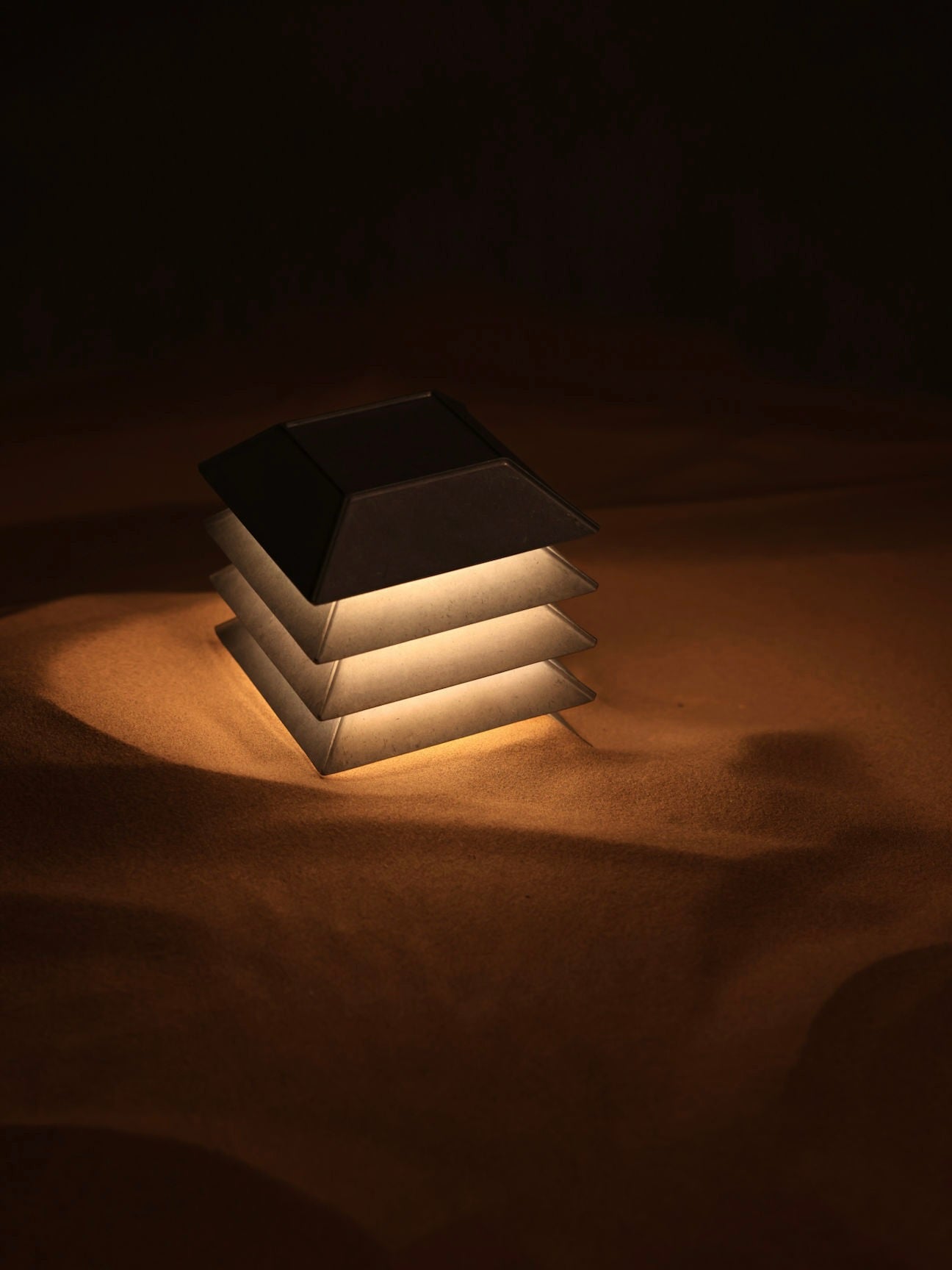 The Pagoda Table Lamp by FEBRERO SHOP casts a warm glow, beautifully lighting up the surrounding sandy surface in the dark with its modern design.