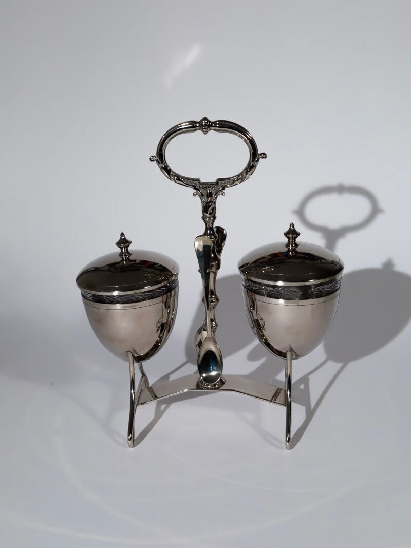 The Vintage mid-century serving set by Break The Mould Magasin boasts a silver double condiment holder with ornate handles and two lidded bowls on a stand. The polished bowls elegantly reflect light, casting subtle shadows on a plain white background, accompanied by gleaming silver-plated spoons and accented with chrome highlights.