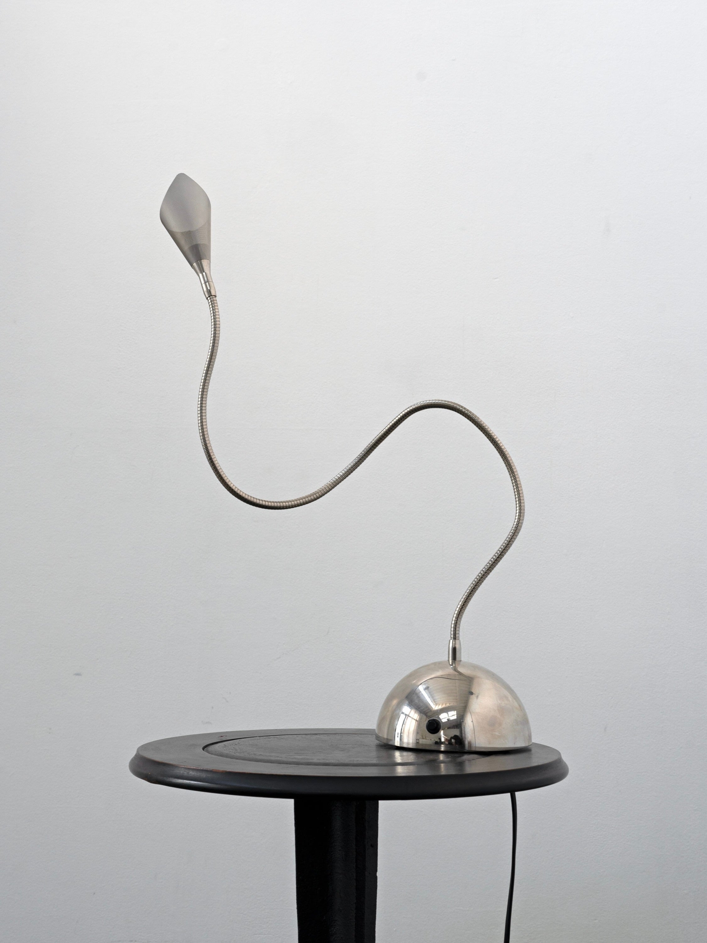 The Italian Chrome Table Lamp, c.1990 by Tom Bogle, with a flexible neck and dome base, stands on a round black pedestal table against a light gray wall, embodying the refined elegance of Italian lighting design.