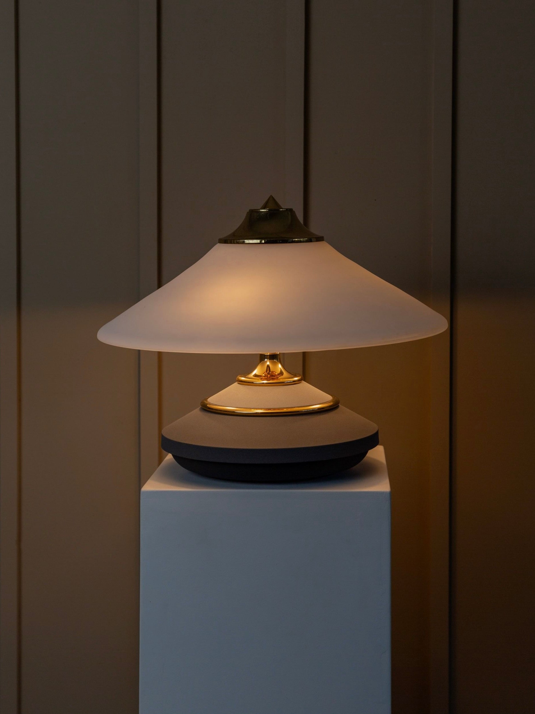 The Memphis Style Vintage Murano Glass Table Lamp by Bottega Jacobs, featuring a conical white shade with a gold accent, stands on a white pedestal. Set against a dark, vertical-paneled background, it casts a warm glow that evokes the elegant charm of the 1980s.