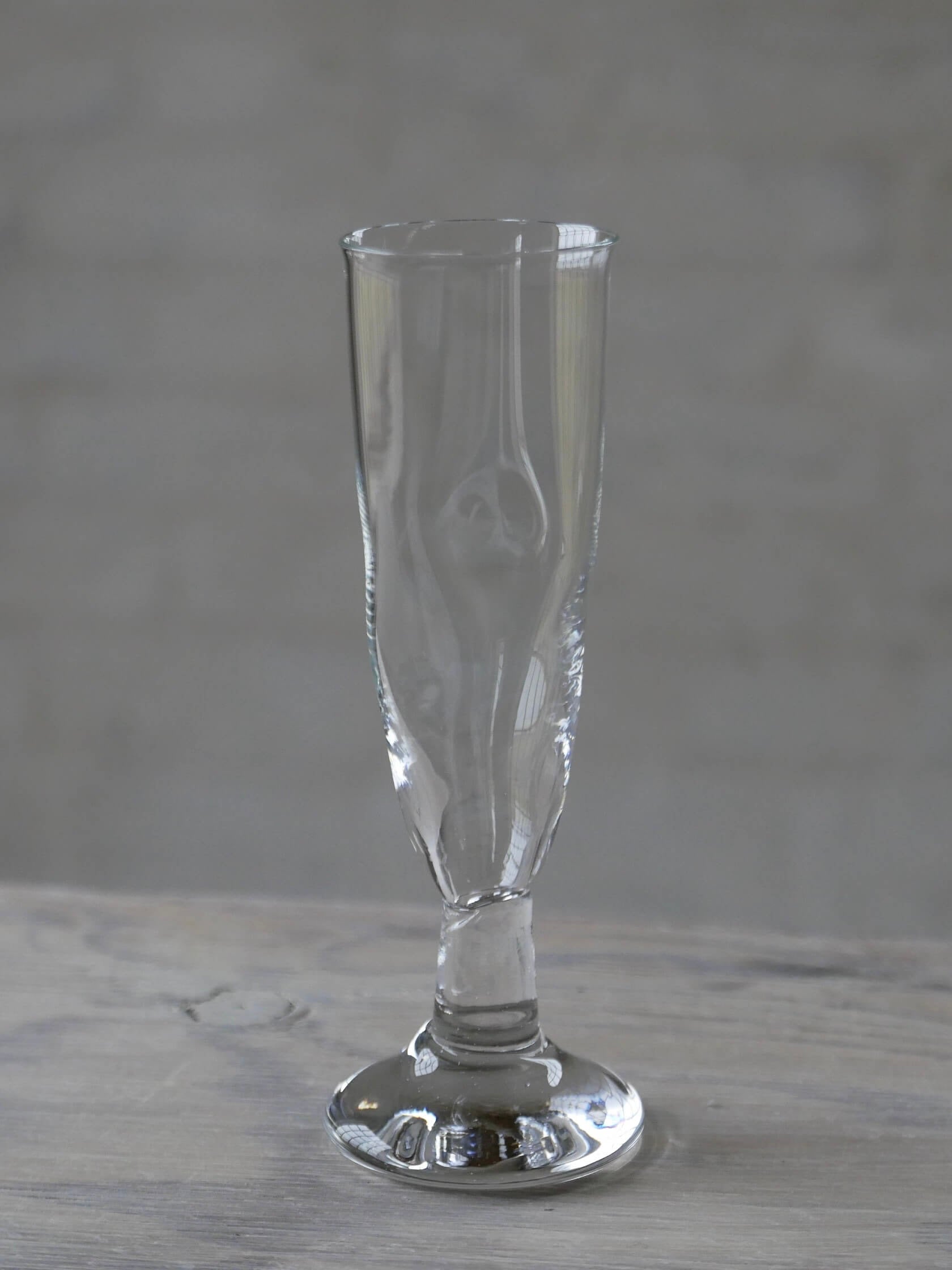 A tall Twisted Flute from Silje Lindrup, with a clear body and narrow stem and base, rests on a wooden surface. The elegant glassware appears slender and is empty, with subtle reflections on its surface. The background is blurred and grey.