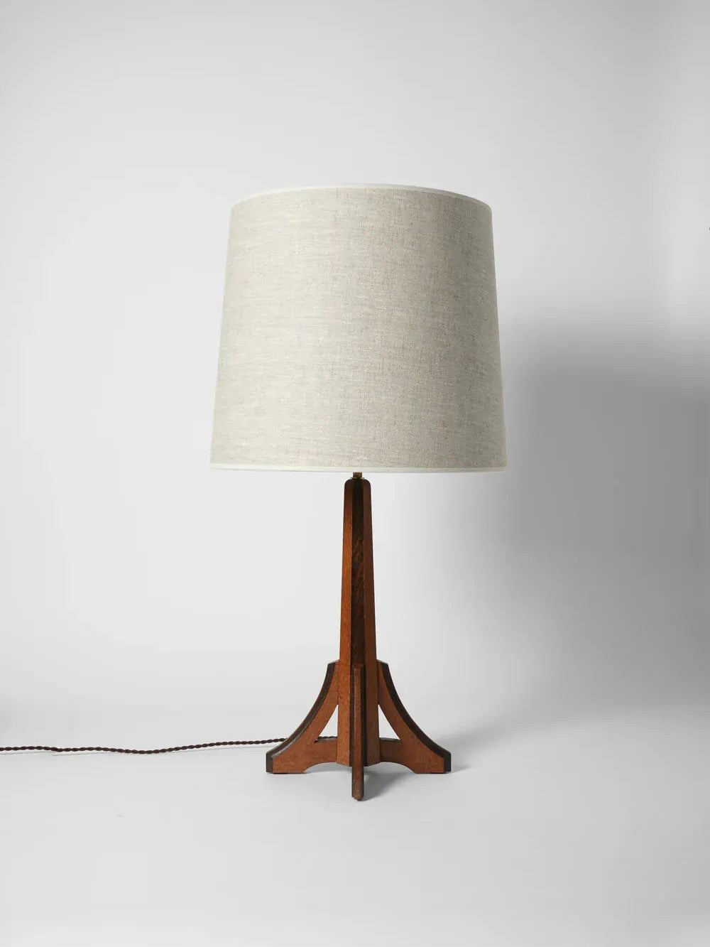 The Arts & Crafts Oak Lamp from the 1920s, designed by Barracuda Interiors, showcases a bicolor oak frame with a light grey fabric lampshade and a wooden triangular base, standing elegantly against a plain white background.