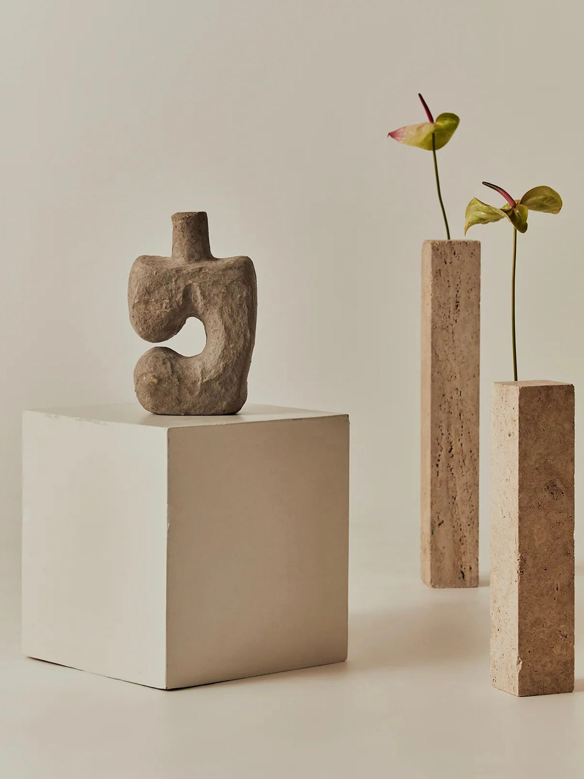 A modern art scene showcases a stone sculpture on the Chantada Side Table by MarlotBaus, flanked by tall rectangular vases with pink anthuriums. This setup is set against a minimalist beige backdrop, seamlessly blending contemporary design with versatile furniture elements.