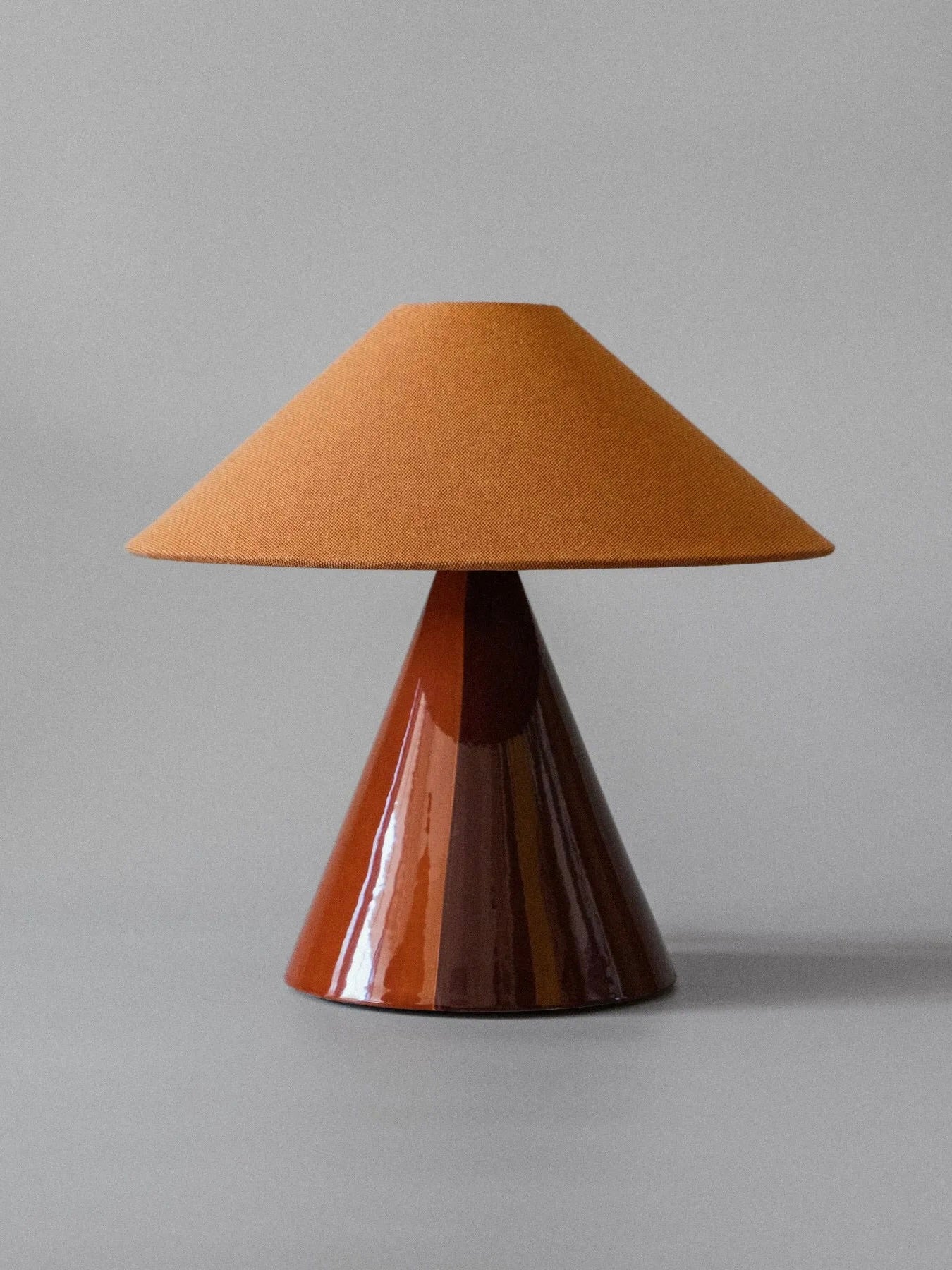 The "Caterina" Table Lamp in Cognac by mattina moderna, featuring a brown cone-shaped base and a wide conical fabric shade with an elegant hand-painted design, is placed on a gray surface against a light gray background.