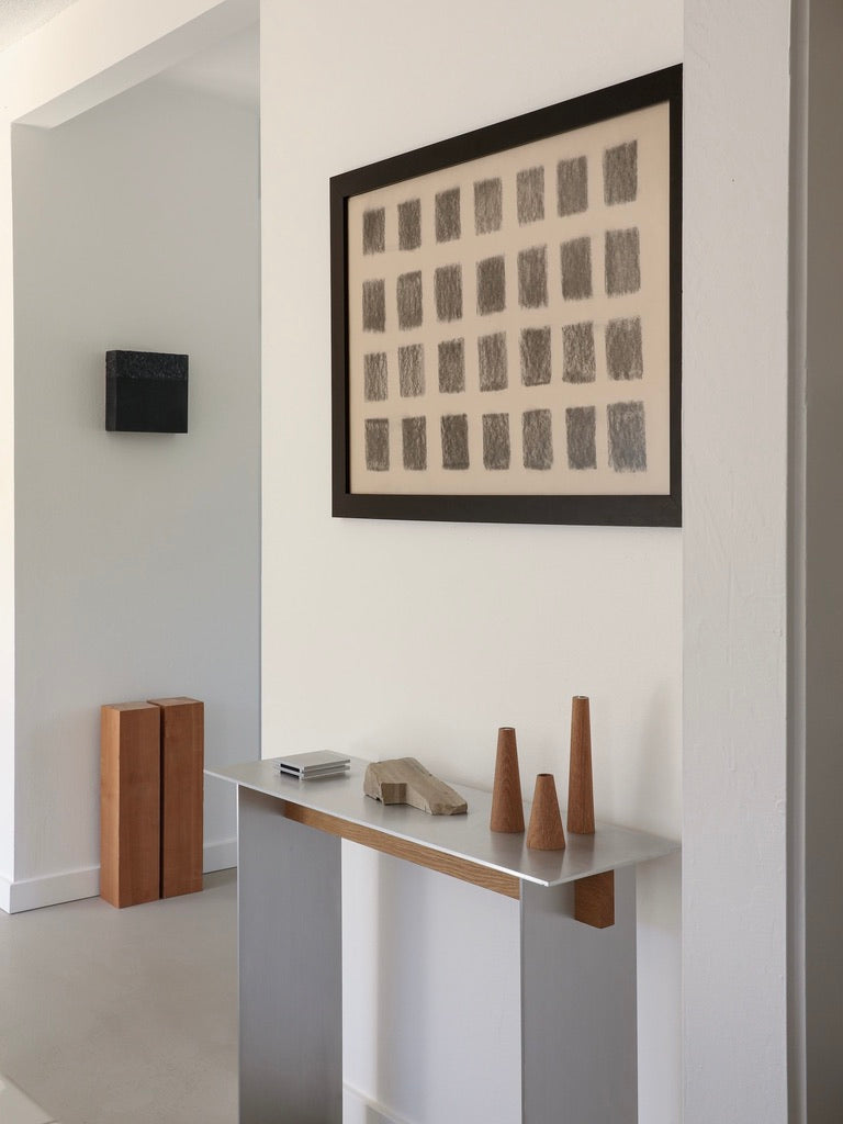 A minimalist interior showcases the Studio Orlando Pippig AL-02 Console Table with wooden geometric sculptures. An aluminum furniture touch is present, alongside a large artwork of dark rectangles. A wooden sculpture and small black artwork complete the look.
