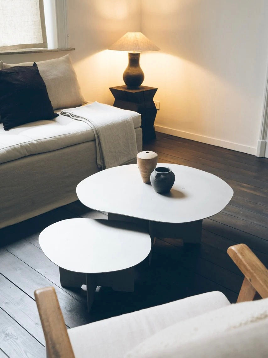 A cozy living room featuring a beige sofa with dark brown and beige cushions, a wooden armchair, and KØGE Design's Double Shape Coffee Table set in white, crafted with sustainable manufacturing. A sculptural black lamp with a beige shade illuminates the space from an end table beside the sofa.