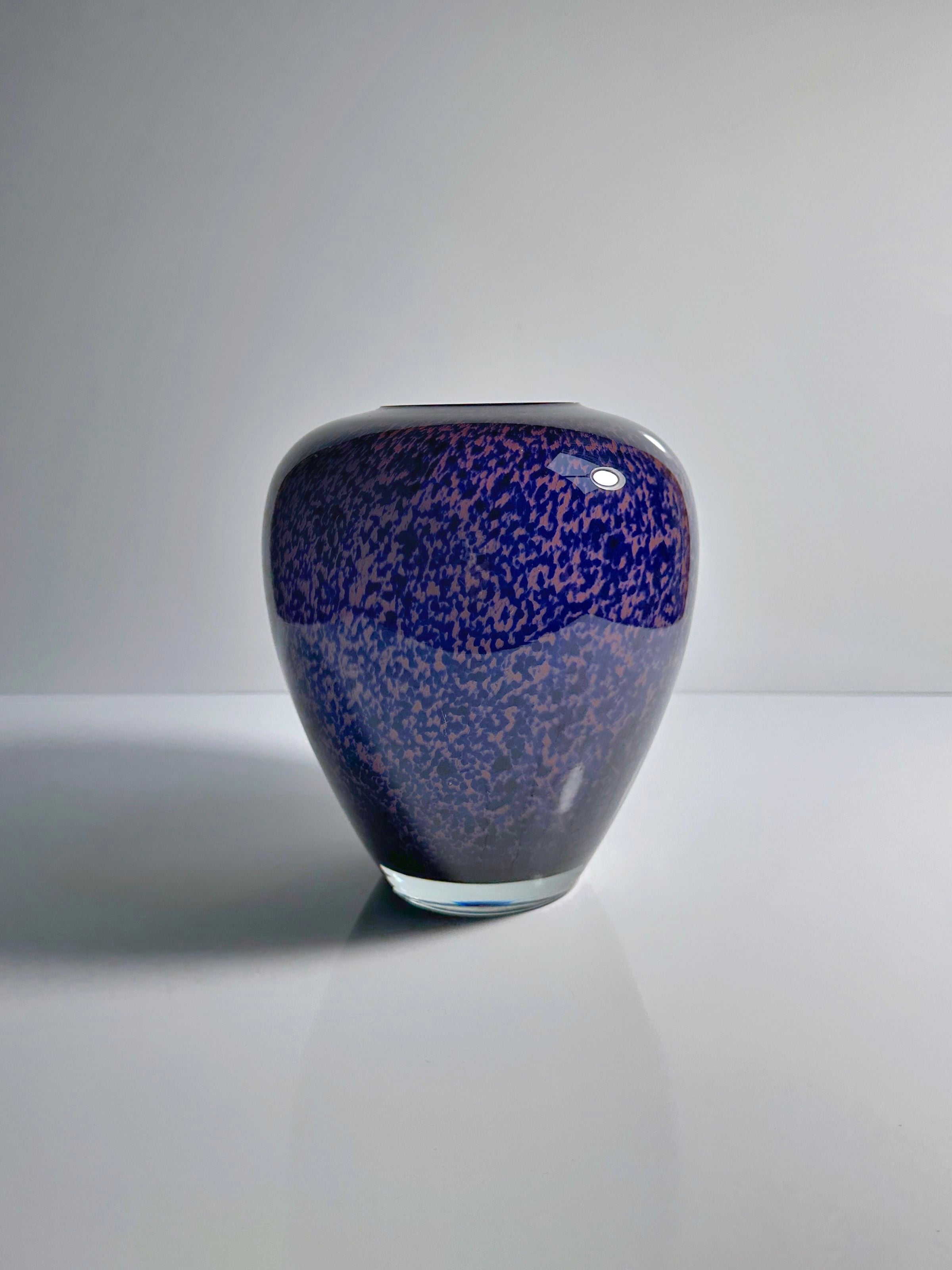 The 1930s Art Deco Tortoise Glass Vase by Dodo Vintage, with its speckled purple and blue pattern, sits elegantly on a smooth white surface against a light gray background, exuding the timeless charm of a collector's item.