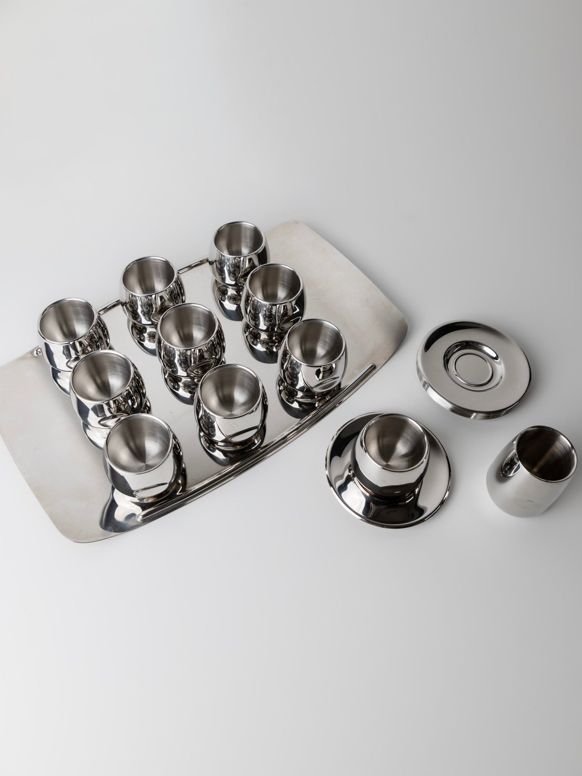 The "Coffee set by AMC 70s" from Bottega Jacobs shines with a minimalist design, featuring eight small cups on a rectangular tray and two teapots with lids on the right, all in polished silver. The sleek metal reflects light beautifully against the white surface.