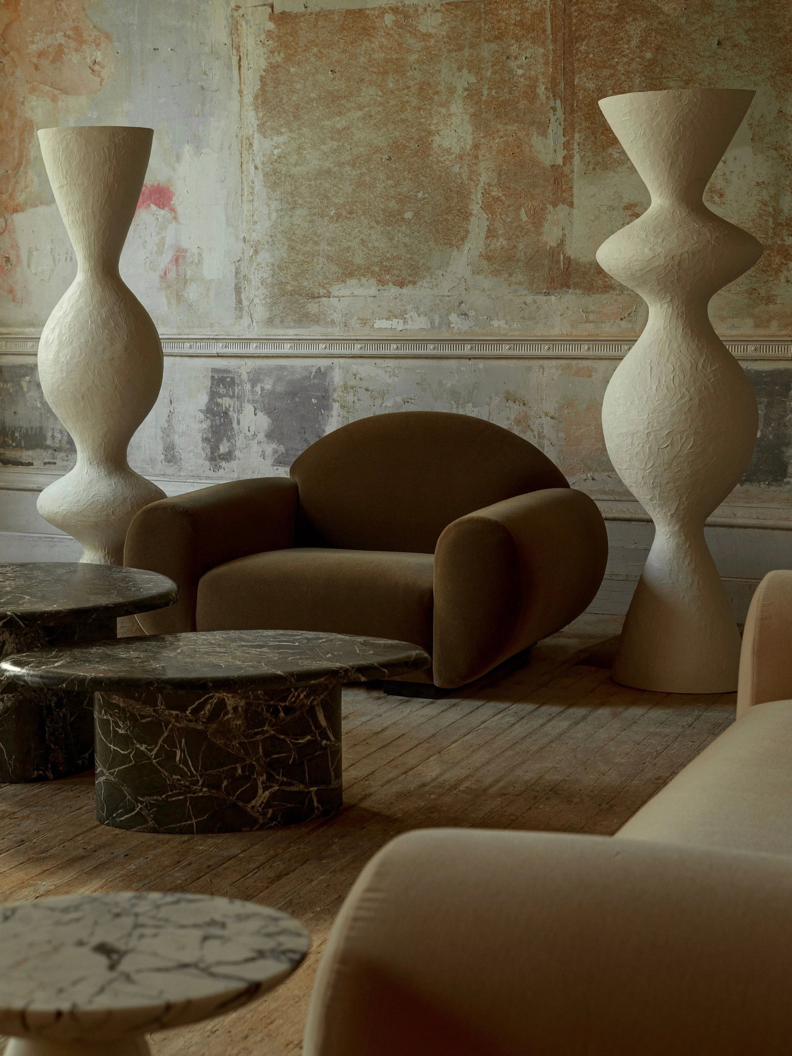A room with textured walls showcases modern furniture, including curved sofas upholstered in luxurious mohair velvet and round marble tables. Two tall, sculptural white lamps introduce a distinctive design element. The setting, enhanced by the bold and sensual "Sparta" Armchair from Ransom & Dunn, features a warm, earthy color palette.