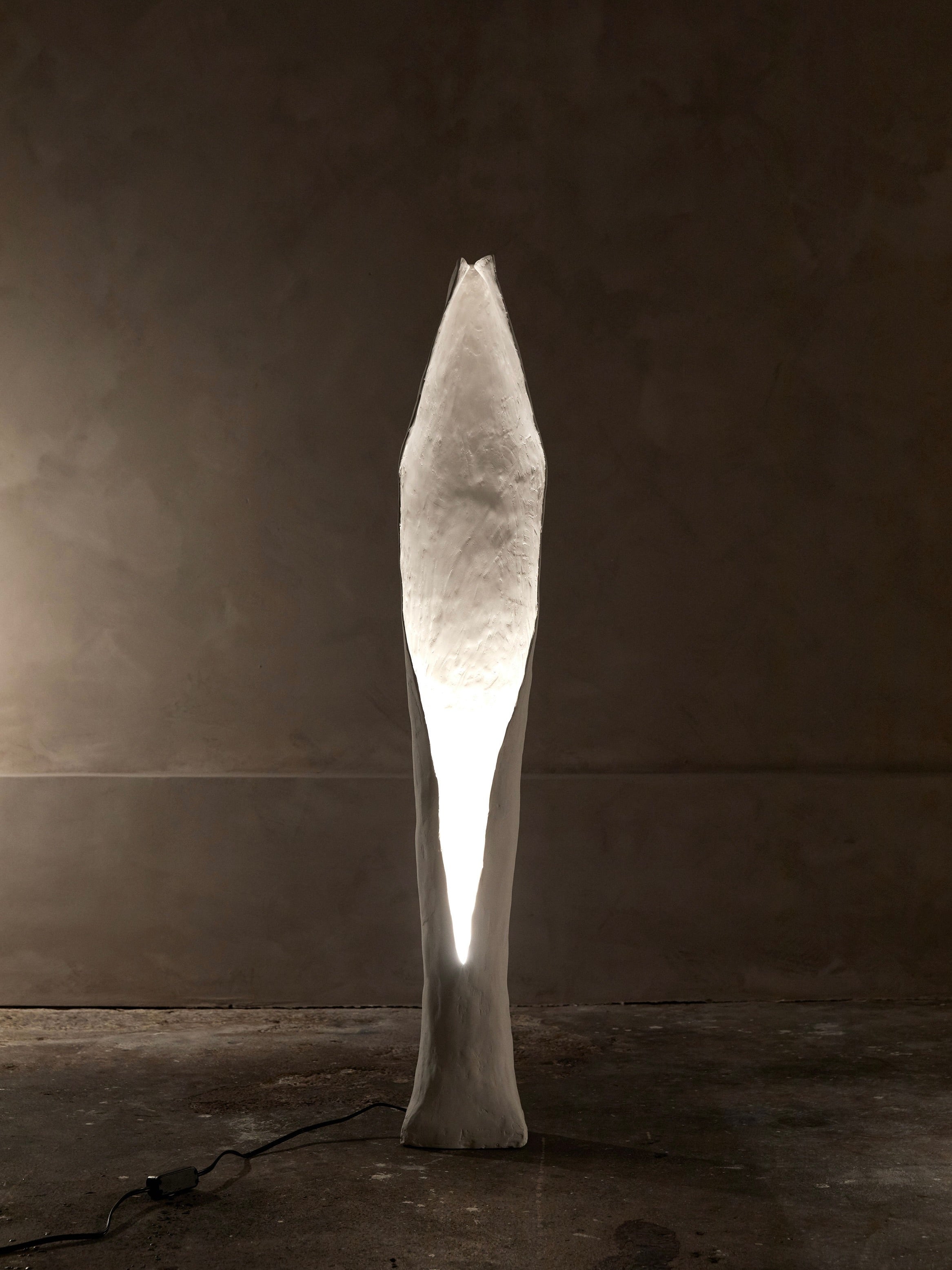 A tall, abstract sculpture with a narrow oval-shaped light is reminiscent of the white and textured Alcove Floor Lamp by Studio Tristan Kallas, standing on concrete against a dimly lit, neutral background.