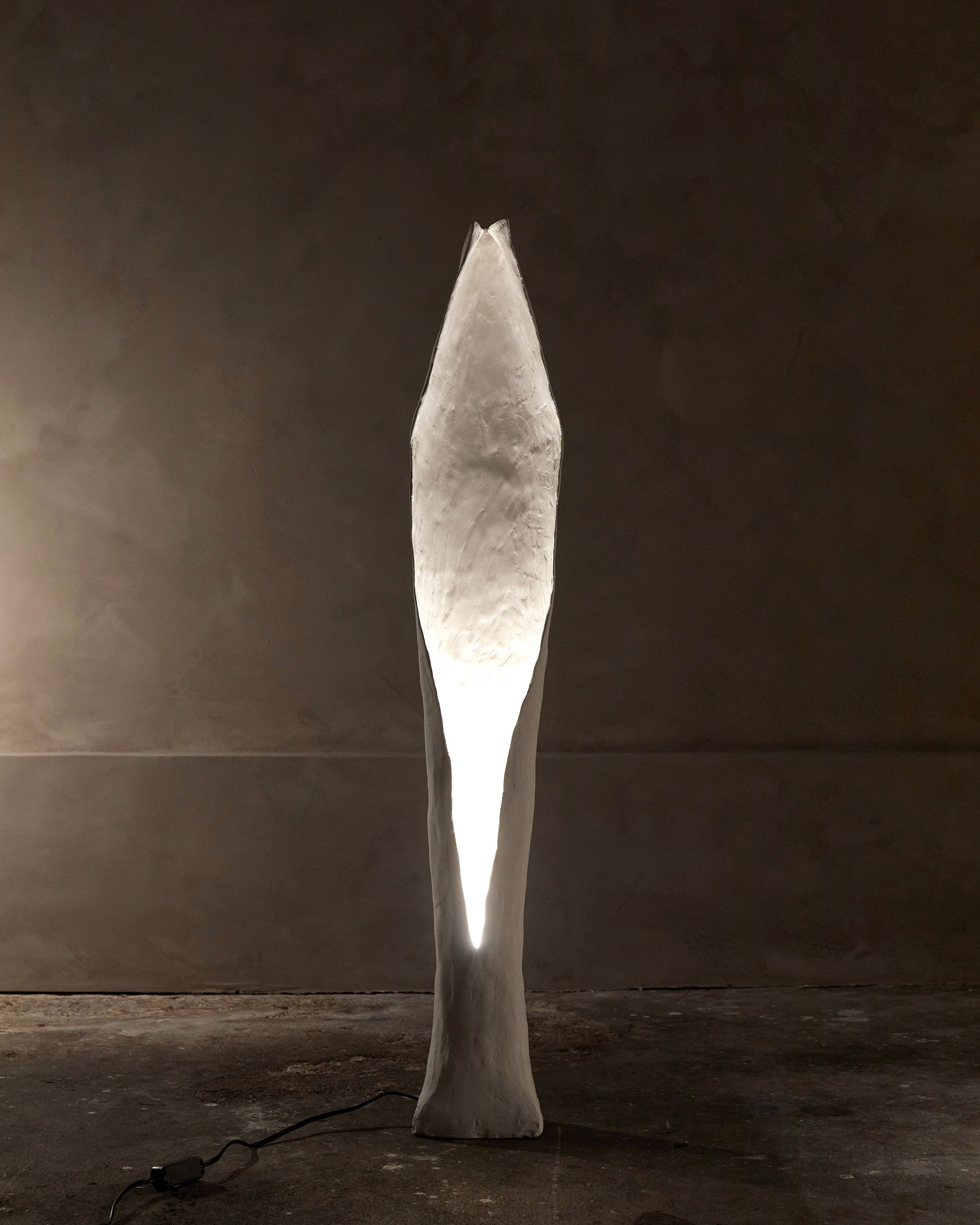 A tall, abstract sculpture with a narrow oval-shaped light is reminiscent of the white and textured Alcove Floor Lamp by Studio Tristan Kallas, standing on concrete against a dimly lit, neutral background.