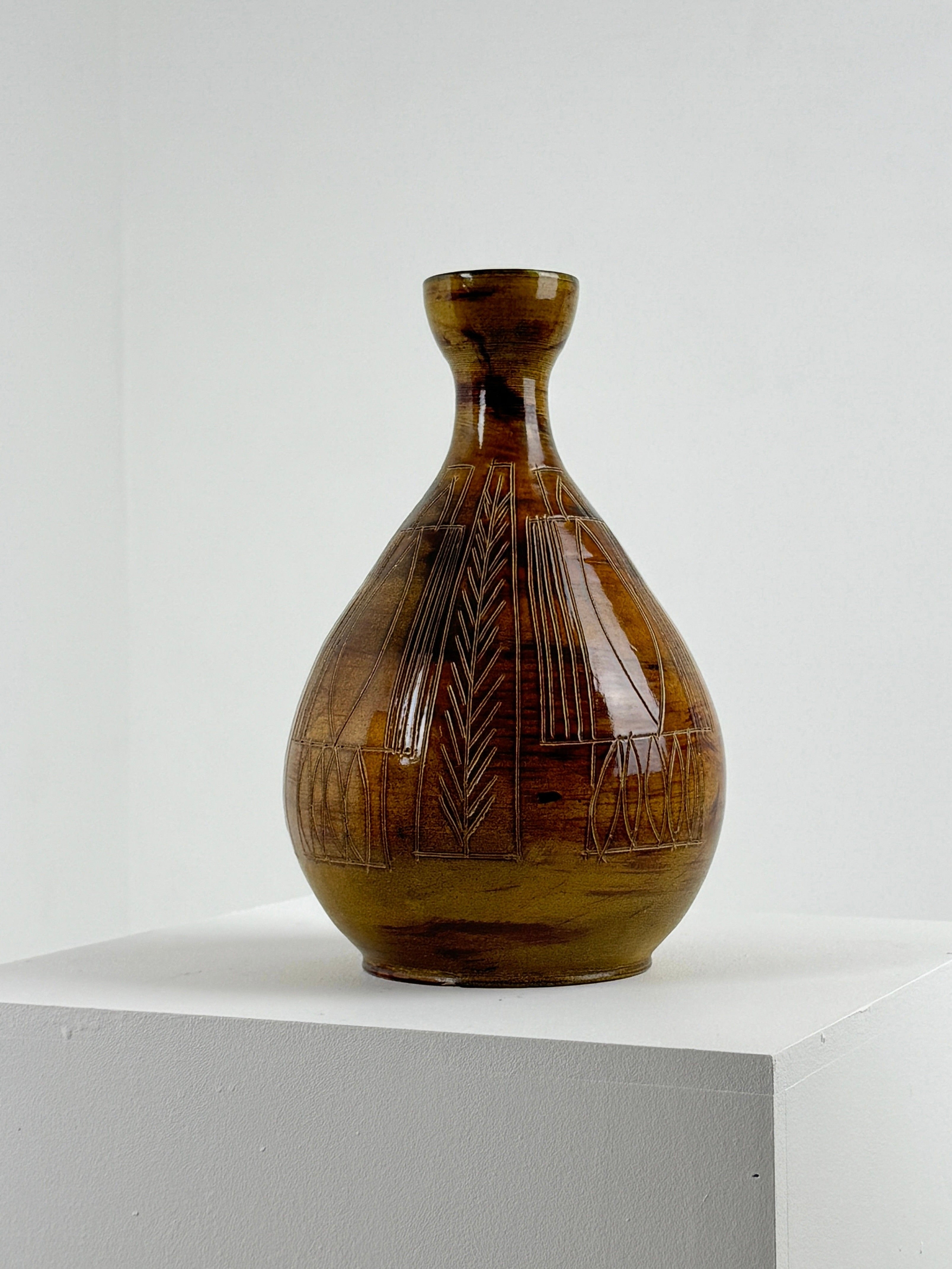 A unique Ceramic Vase by the potters of Accolay, offered by Figuresdesign, with a polished brown finish and incised geometric decoration featuring vertical lines and leaf motifs, is displayed on a white pedestal against a plain background. This exquisite piece showcases a narrow neck and rounded body.