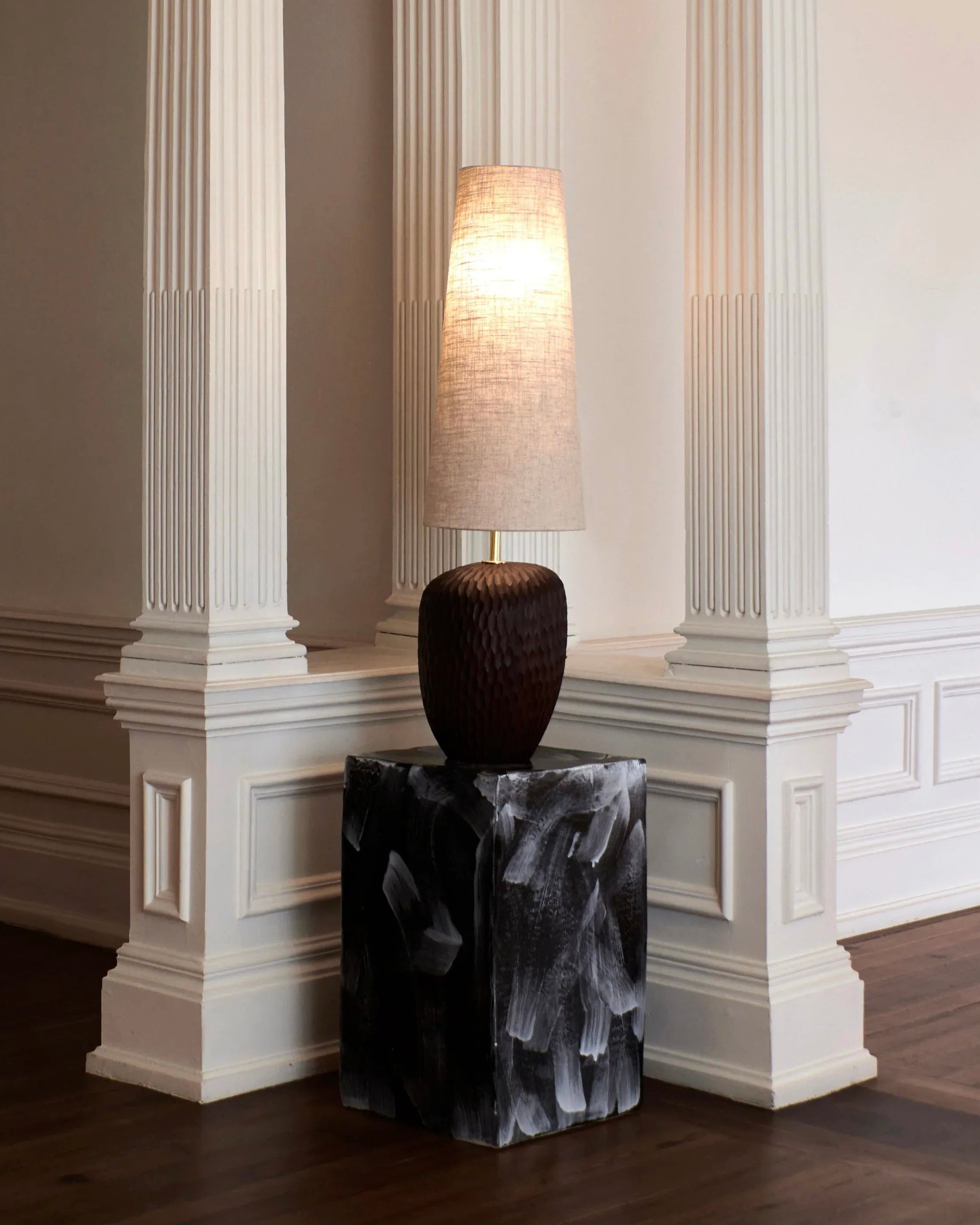 A tall fabric lampshade atop a textured dark brown base is placed on the Ceramic Side Table with a geometric shape from Project 213A. This setup is positioned between two white, ornately carved pillars in a minimally decorated, elegant room with wooden floors, adding modern flair.