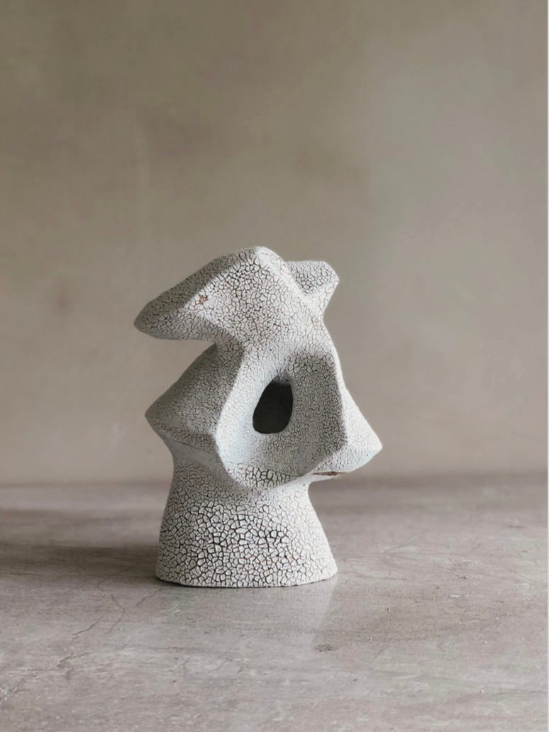 The 'Visions of Light' sculpture by Emma Lindegaard Studio is an abstract ceramic piece with a textured, cracked surface. Crafted with geometric shapes and organic curves featuring a central hole, its tactile finishes stand out against a neutral background on a smooth surface.