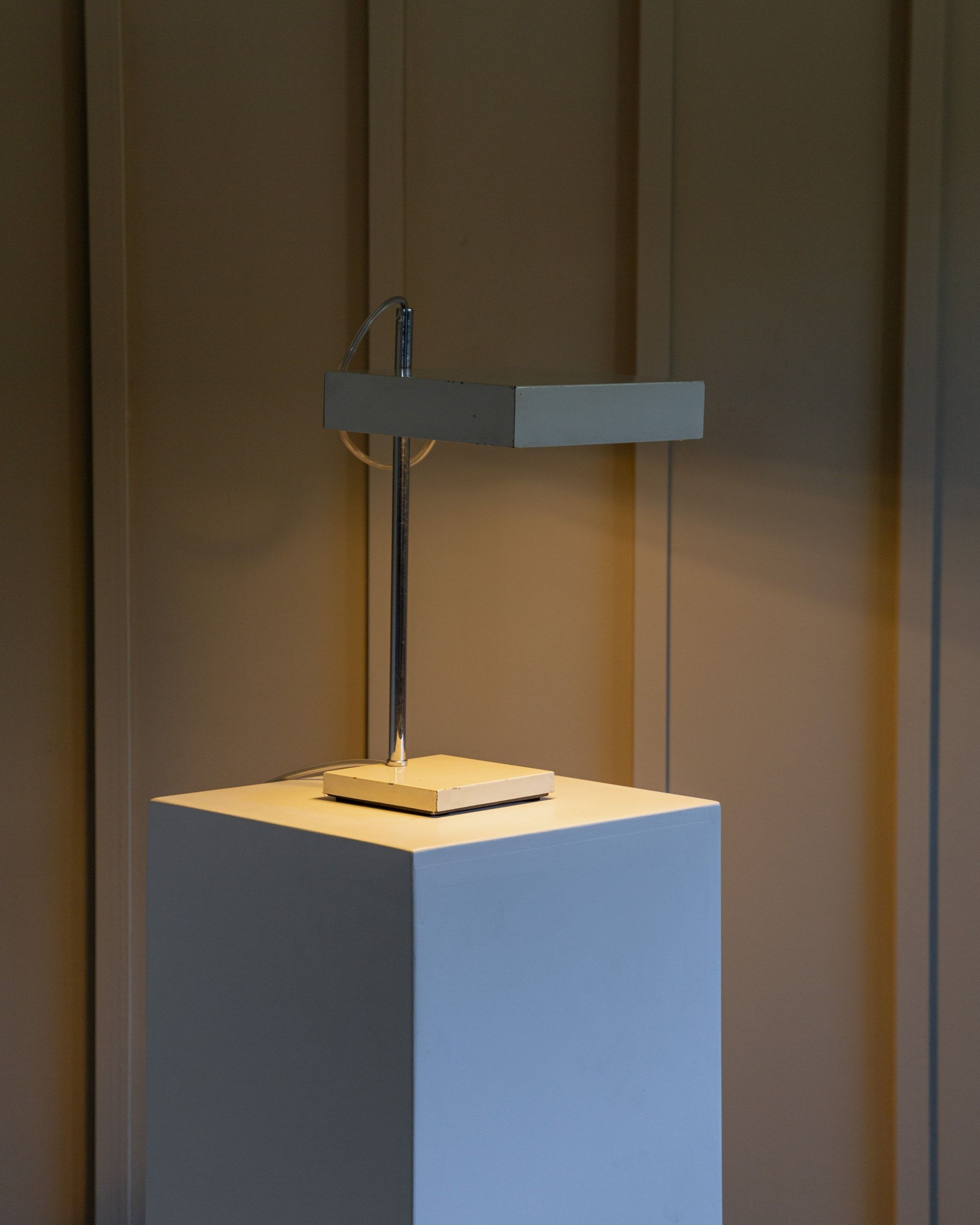 The Kaiser Leuchten Table Lamp by Klaus Hempel from Bottega Jacobs, featuring a sleek rectangular design, is elegantly displayed on a white pedestal. The lamp casts a warm light against the backdrop's subtly colored vertical lines on the textured wall, creating a gentle glow that embodies mid-century modern elegance.