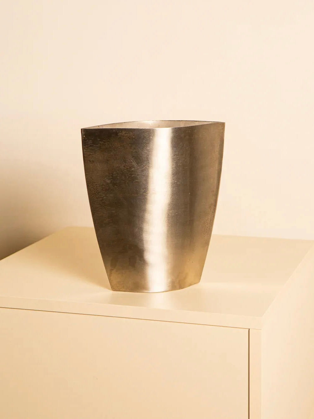 The Treaptyque 80's Italian Metal Vase, with its modern tapered design and shiny metallic finish, sits on a light-colored minimalist cabinet. A softly lit neutral background highlights its smooth surface and reflective qualities, echoing 1980s elegance and craftsmanship.