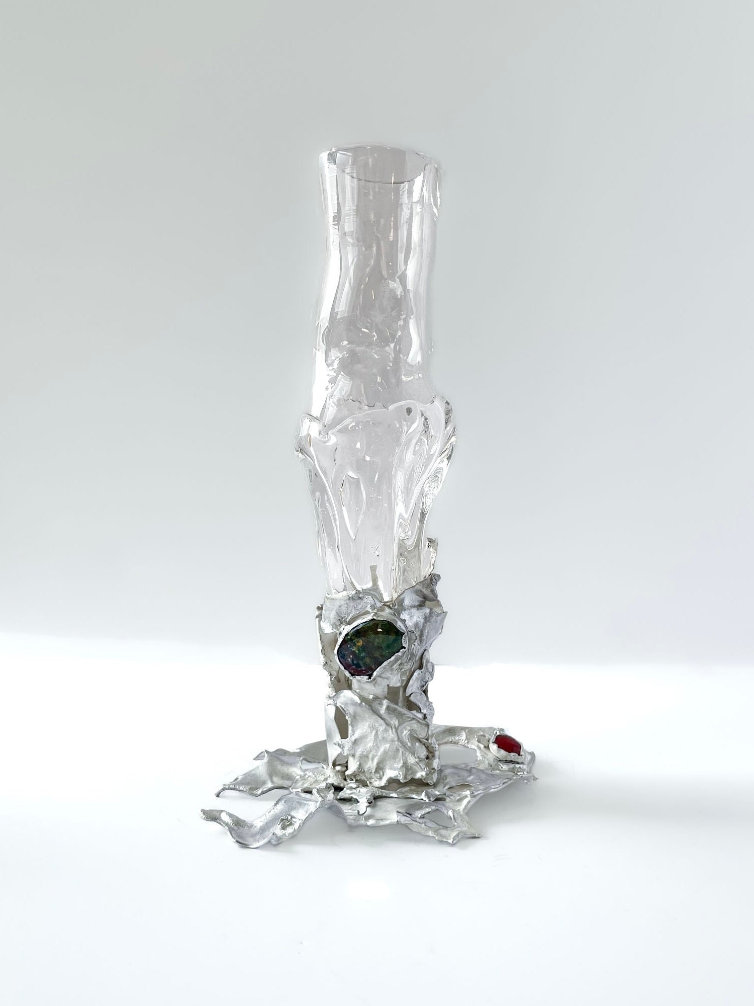 The Jewelled Goblet no. 7 by Lindsey Fontijn from Szkło Studio is a tall, abstract hand-blown glass sculpture with a smooth, transparent upper portion and an irregular metallic base. The base features colorful embedded natural gemstones, prominently including a green one. The piece rests on a white surface with a light background.