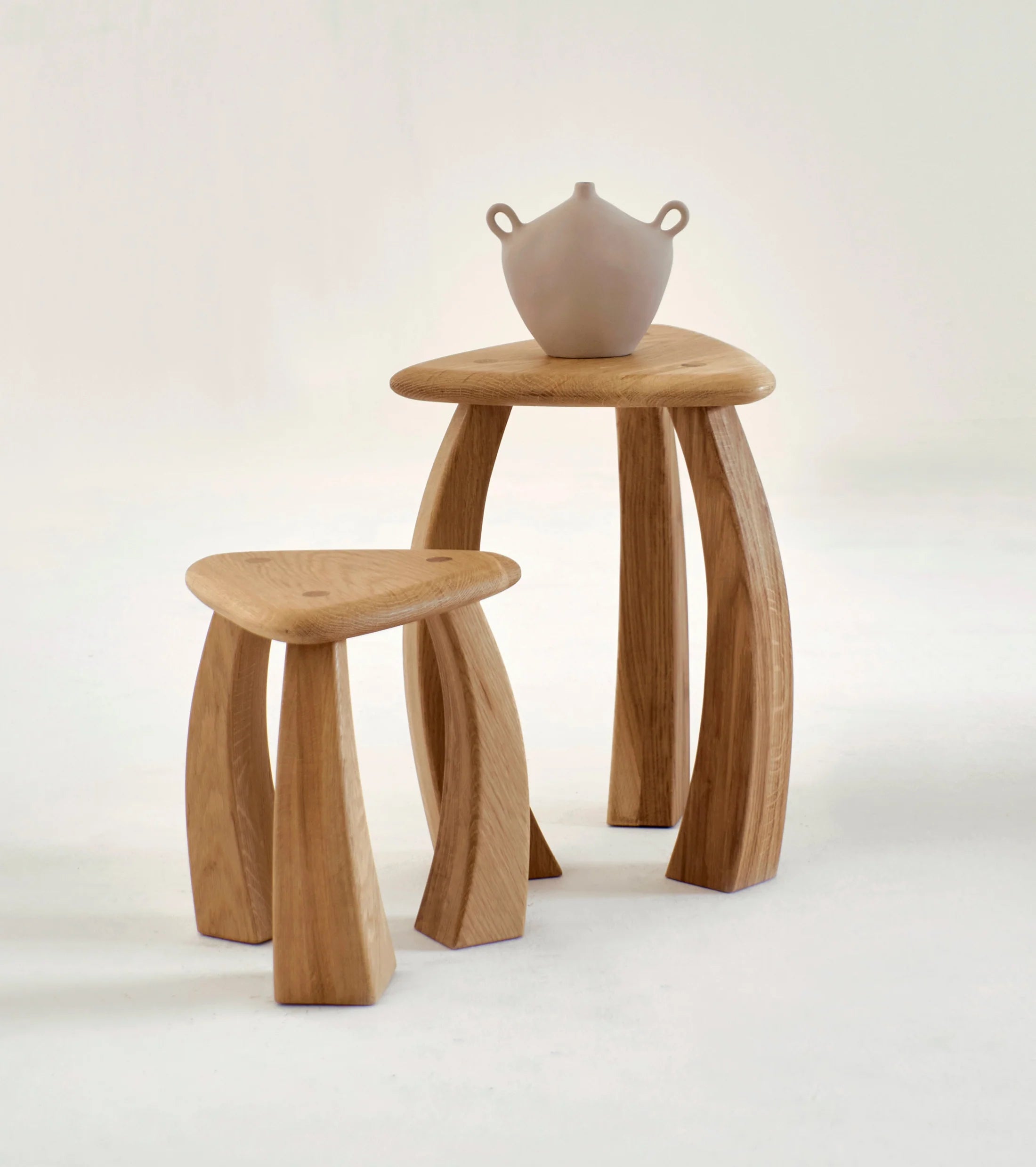 Two Arc de Stool '37 wooden stools from Project 213A with curved legs are placed on a white surface. The taller stool has a smooth jug with two handles on top. These stools feature a natural wood finish and embody a modern, minimalist design that blends functionality with aesthetic appeal, making them perfect for versatile seating in any modern home décor.