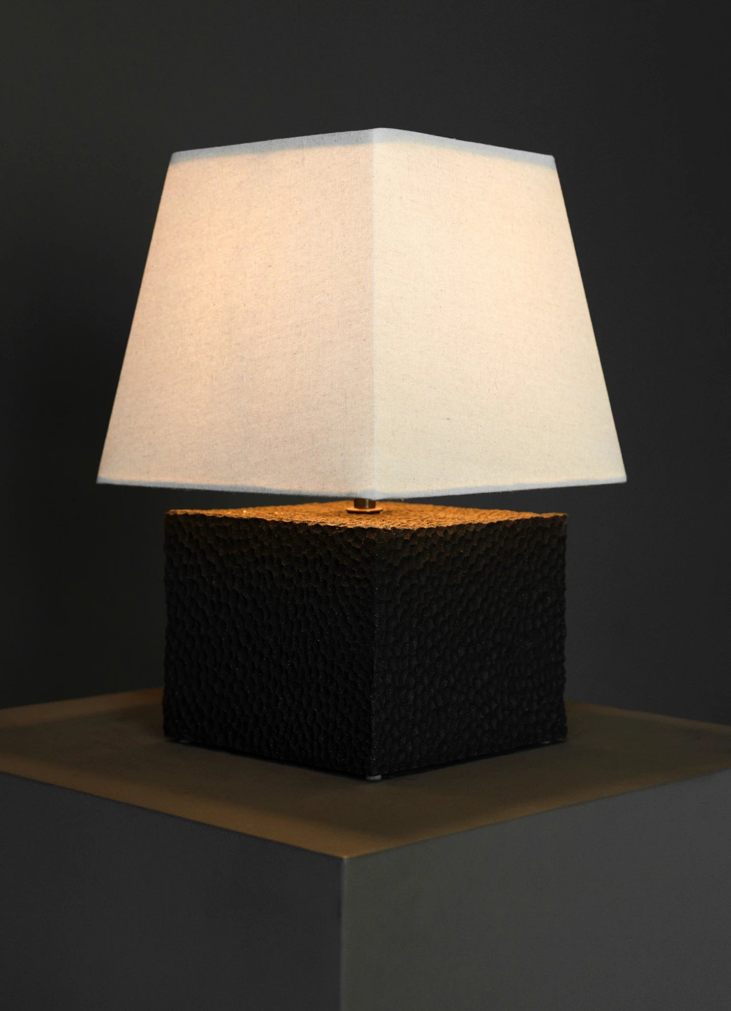 The Akune Ceramic Lamp - Number Five by Studio OSKLO, featuring a white square lampshade and a textured black square base inspired by post-Bauhaus brutalism, sits illuminated on a dark surface against a dark gray background.