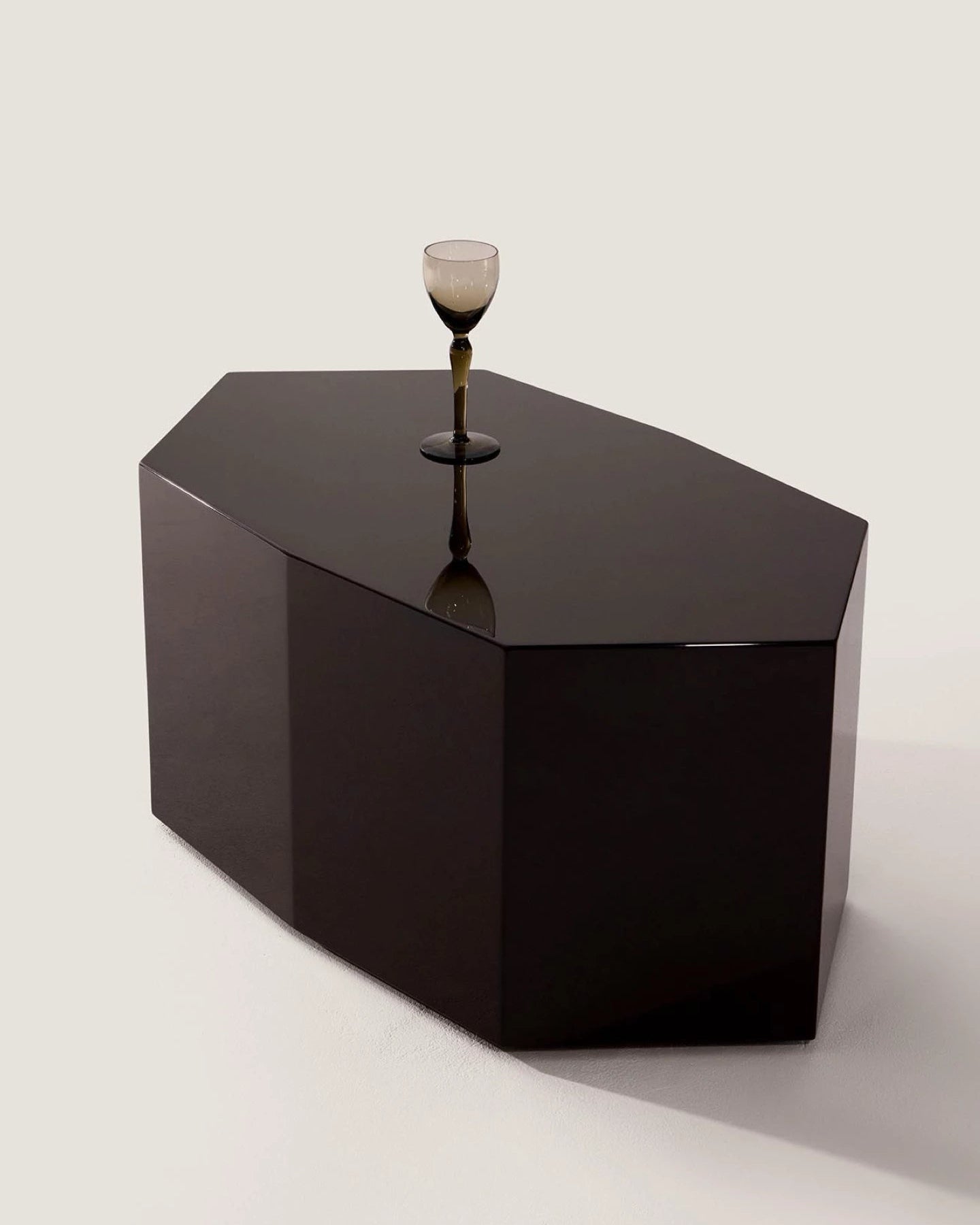 The 'Ame III' Lacquer Coffee Table by Zoé Wolker Studio, with its bold geometric design and glossy dark finish, stands on a light surface. A single glass with a dark stem sits at the center, evoking collectible artistry.
